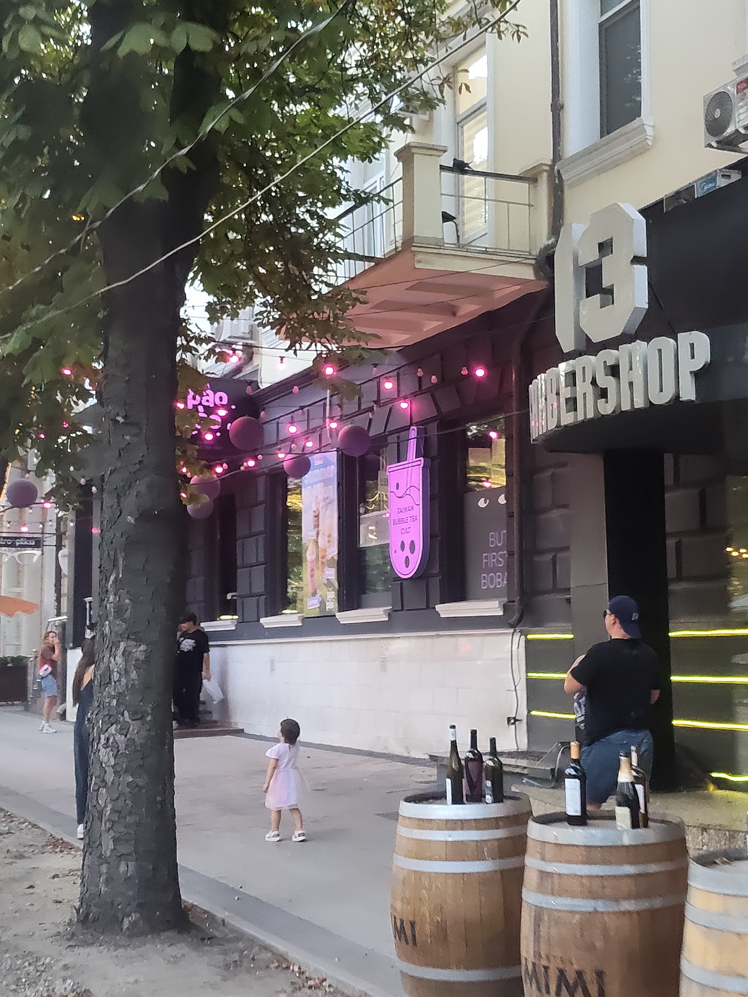 The image depicts a street scene in Chișinău, Moldova, featuring a building with a barber shop sign. A large tree with green leaves is prominent in the foreground on the left, with strings of pink lights and spherical purple decorations hanging from its branches. The barber shop, labeled "13 BARBERSHOP," has a dark facade with visible signage. Next to it, a neon sign in the shape of a bubble tea cup reads "TAIWAN BUBBLE TEA CULT." There are several people in the image: a young child in a light-colored dress stands in the middle of the sidewalk, facing away from the camera; a woman in a dark shirt and jeans stands near the child; and a person in a dark shirt and blue shorts with a blue cap is seen from the back, near the barber shop entrance. In the lower right corner, three wooden barrels are visible, with several bottles of wine and champagne placed on top of them. The street is paved with concrete, and there is a curb separating the sidewalk from a surface with fallen leaves.
