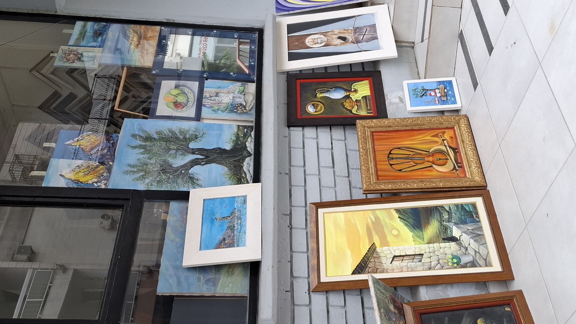 The image shows an outdoor display of framed paintings in Budva, Montenegro. The paintings are leaning against a light gray brick wall and a window, some partially obscured by the window frame and reflection. The paintings depict a variety of subjects, including landscapes, still lifes (fruit), a tree, and a musical instrument (violin). The frames vary in style and color. The location is suggested by the architecture and presumably is a street-side art display. No people are visible.