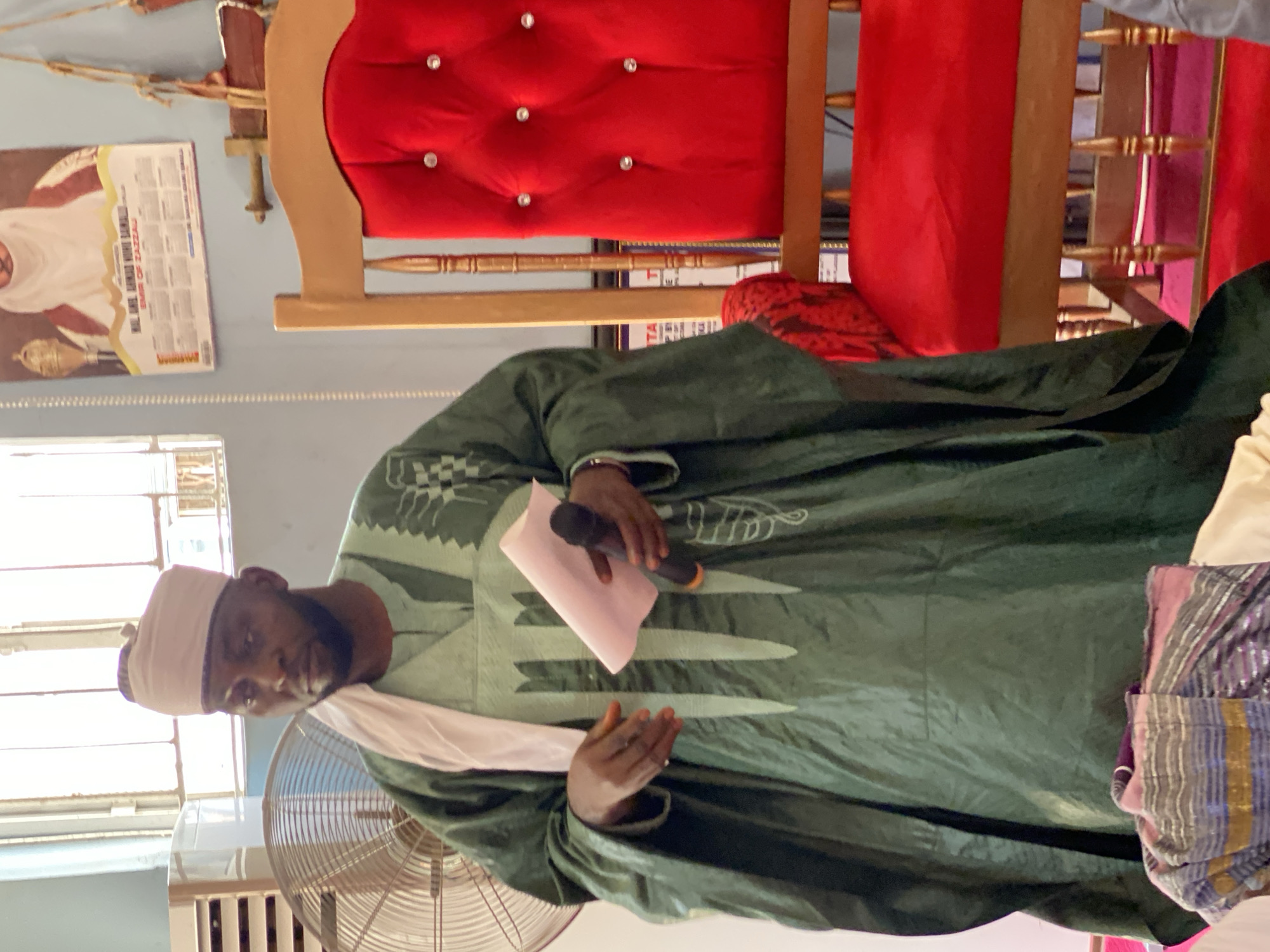A man in a green robe and a white cap is speaking into a microphone while holding a pink paper. He is standing in front of a fan, with a red-cushioned chair behind him. A poster is affixed to the wall in the background, near a window. The location is Sabon Tasha, Nigeria.