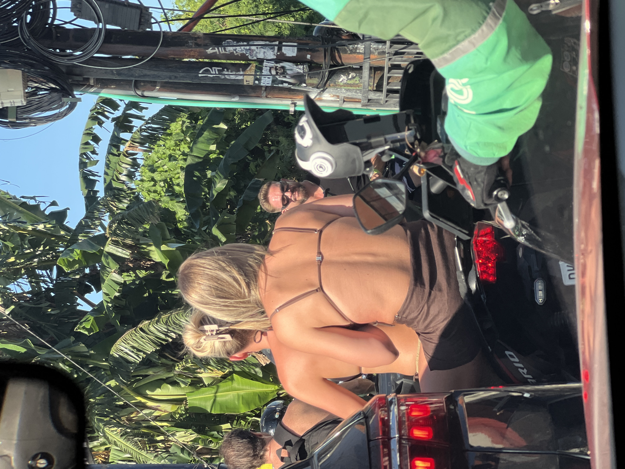 Observed in Unknown, Unknown, is a scene depicting a blonde woman with a hair clip in a bun, possibly in a brown bathing suit or similar attire. She is positioned near a dark-colored vehicle, potentially a car or motorcycle, with visible tail lights. A man with sunglasses is also present in the background, partially obscured. Overhead, there are power lines and lush, green tropical foliage suggesting an urban environment in a tropical or subtropical region. A person in green attire is seen near a motorcycle.