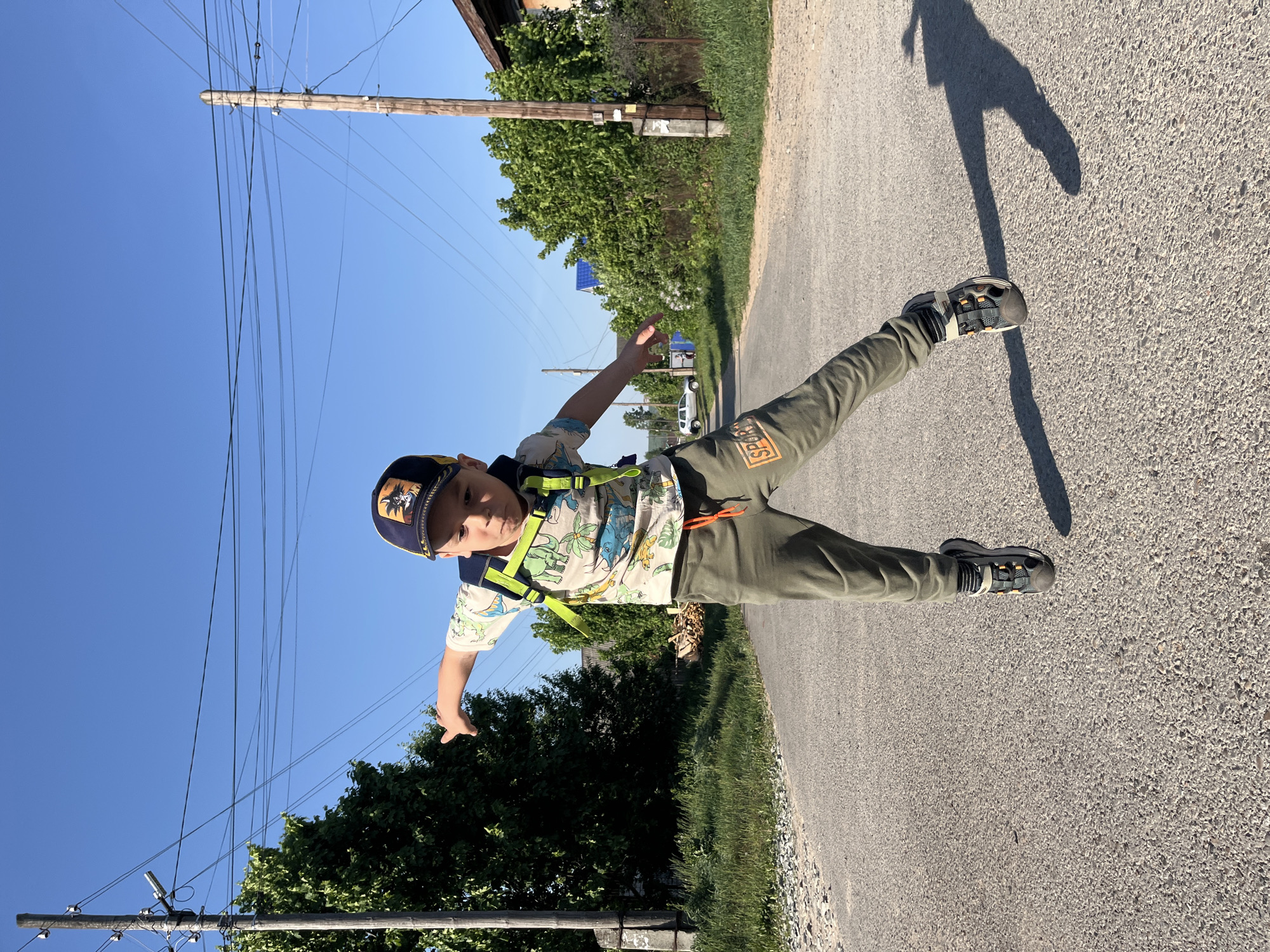 A young boy stands on a paved road, positioned horizontally in the frame. He is wearing a cap, a short-sleeved shirt with a dinosaur pattern, olive-green pants, sandals, and a neon-yellow backpack. He has his arms outstretched, with his legs spread apart. His shadow is visible on the road behind him.
The environment includes overhead power lines to the left, trees on both sides of the road, and a blurred building in the background. A car is visible further down the road. The scene appears to be set in Chaikovsky, Russia, as indicated by the location information.
