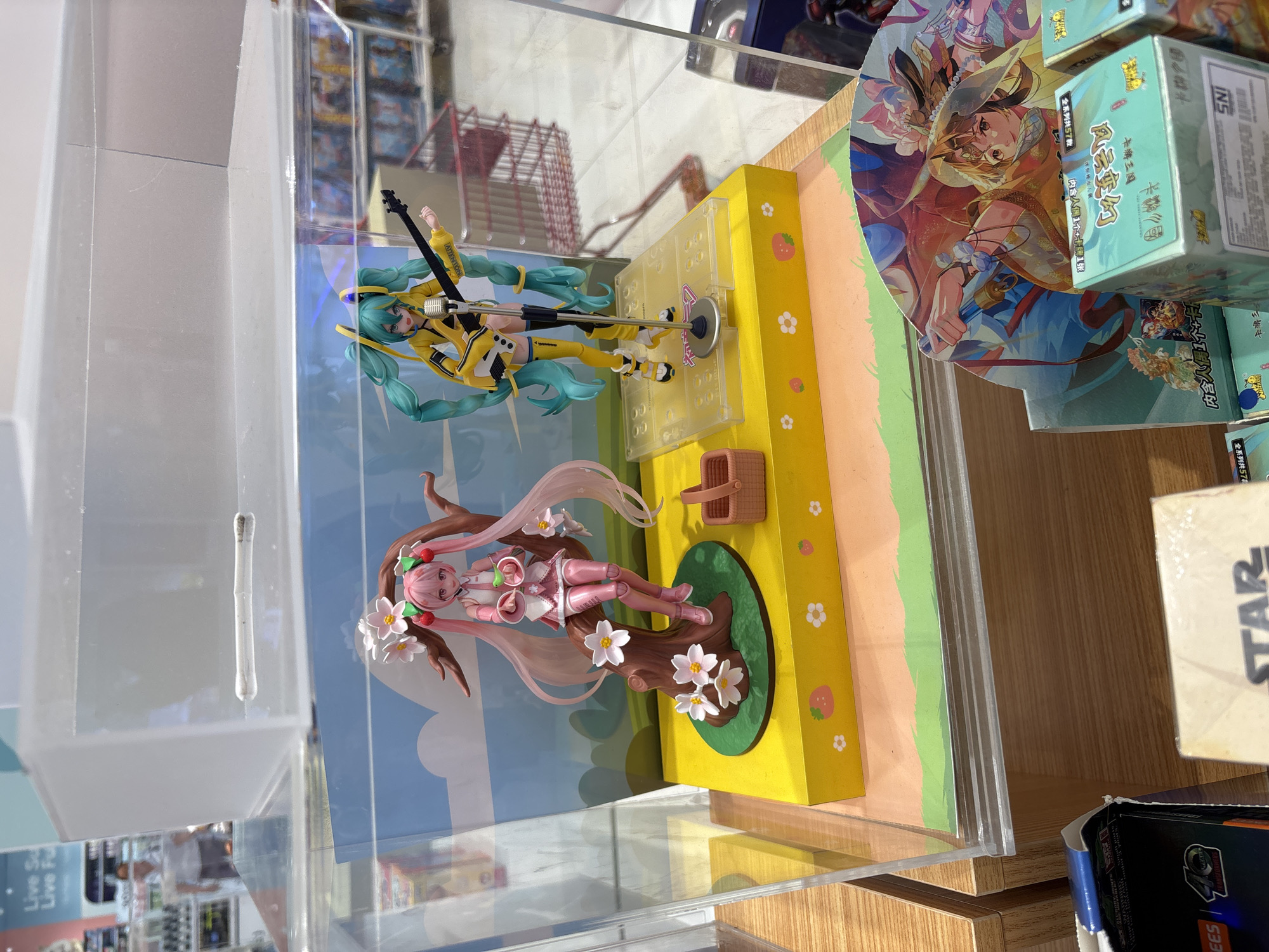 The image shows a display case containing two anime figurines, one with pink hair and a cherry-themed outfit, and another with teal hair and a yellow outfit, holding a guitar. The figurines are placed on a yellow platform decorated with strawberries and flowers. In the background, shelves filled with merchandise are visible, along with a shopping cart and a person in a white shirt. The scene appears to be set in a retail environment, possibly a toy store or a department store. The overall impression is one of a commercial display, highlighting collectible figures. The location is identified as West Jakarta, Indonesia.