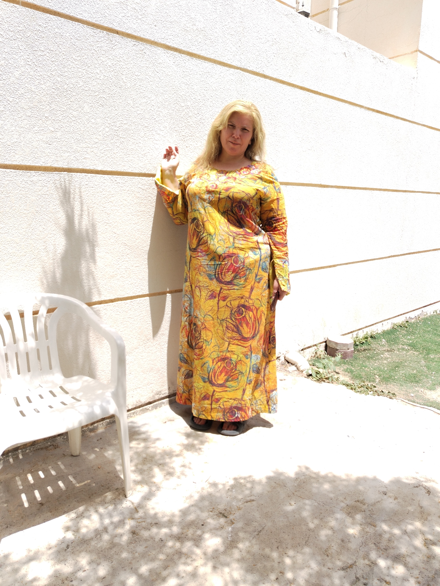 The image depicts a blonde woman in a yellow, floral-patterned, long-sleeved dress standing against a beige stucco wall with horizontal grooves. She has one hand raised slightly. A white plastic chair is located to her left. There is a patch of grass in the background to the right. The ground appears to be a concrete or stone surface dappled with shadows. The location is Al Mas'oudi, United Arab Emirates.