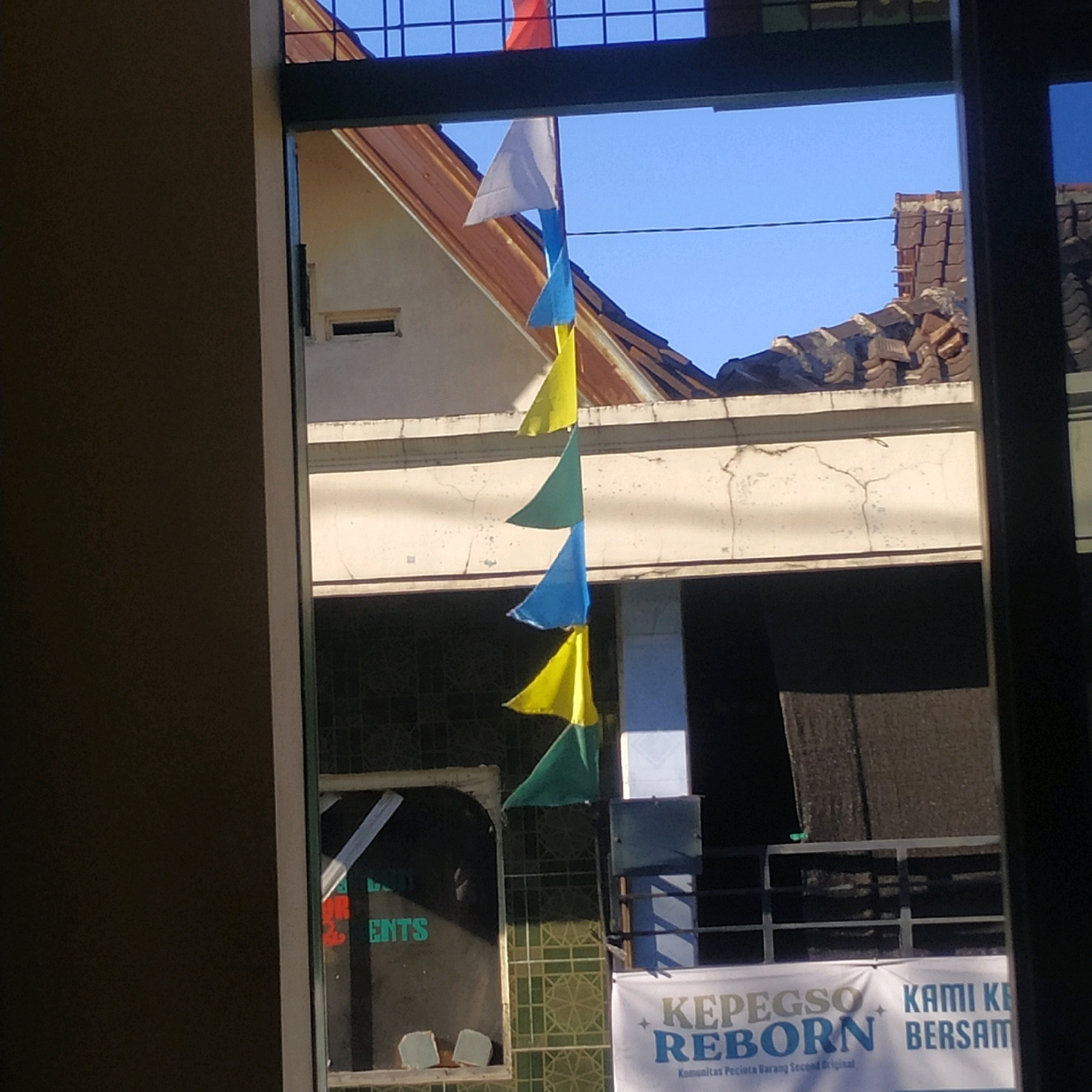 The image captures a scene viewed through a window, with a focus on a string of triangular flags of varying colors—white, blue, yellow, and green—hanging vertically. These flags are silhouetted against a bright blue sky and the tiled roof of a building. In the background, various architectural elements are visible, including roof tiles, a white wall with visible cracks, and what appears to be a window with lettering that includes "ENTS" and "REBORN". The scene suggests an outdoor or semi-outdoor setting, possibly in a place with a clear sky. There are no people or animals clearly discernible in the image. The overall impression is one of a bright, sunny day with some elements of simple decoration, such as the flags, and hints of urban or community infrastructure. The location of Batan, Indonesia, implies a tropical or subtropical climate, which is consistent with the bright sunlight and open sky.
