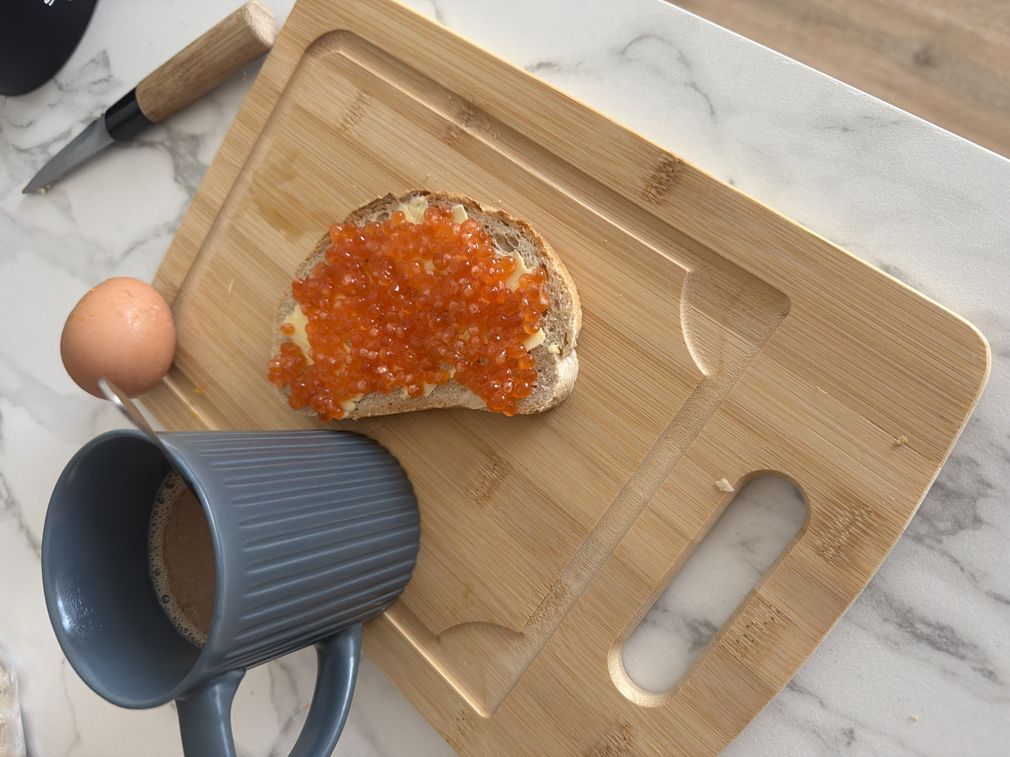 A close-up, overhead shot shows a wooden cutting board with breakfast items on a marble countertop. On the left is a grey, ribbed mug filled with a dark beverage, possibly coffee. A spoon is submerged in the mug. To the right of the mug is a slice of dark bread topped with butter and red caviar. Behind the toast, an uncooked brown egg rests on the cutting board. A knife with a wooden handle lies to the right of the egg. The background is a marble countertop with a wood-grain accent. The scene is well-lit, suggesting it is daytime, likely in a domestic setting. The location is not specified in the image.
