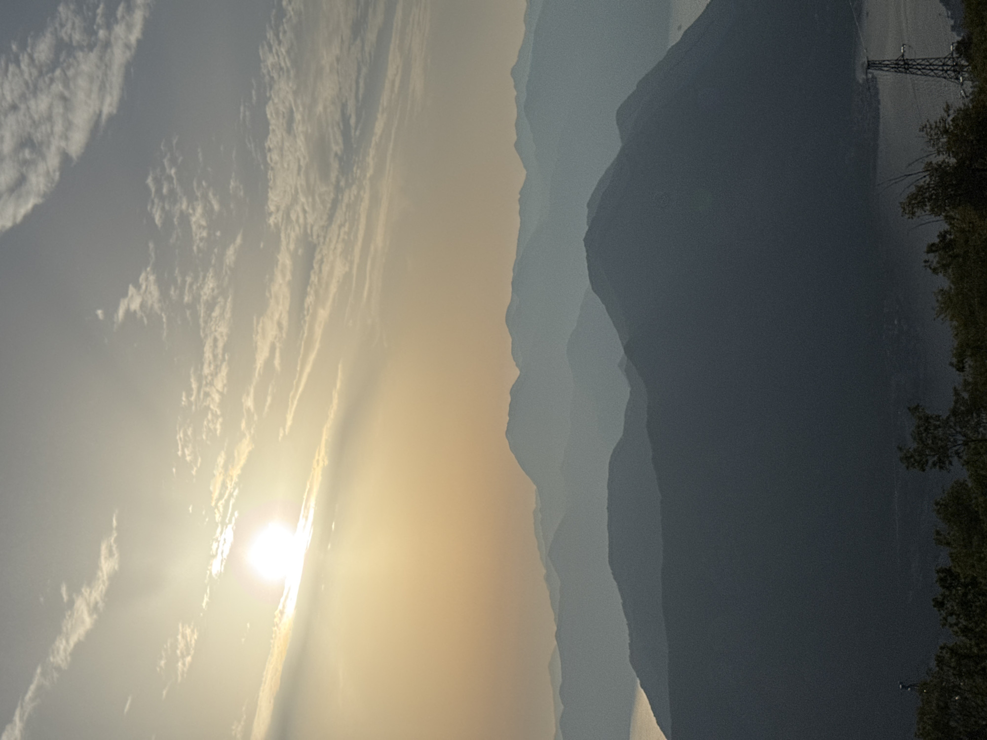 The image captures a panoramic view of layered mountains and a body of water, likely the Bay of Kotor, at sunset, as seen from a high vantage point in Žanjev Do, Montenegro. The sun, low on the horizon and partially obscured by clouds, casts a warm, golden glow across the sky, creating a hazy atmosphere. The foreground features the dark silhouettes of trees and vegetation, with a prominent pylon or tower visible to the right. In the distance, successive ranges of mountains fade into shades of blue and gray, with the shimmering surface of the water appearing between them. The scene is characterized by its serene and majestic natural beauty.