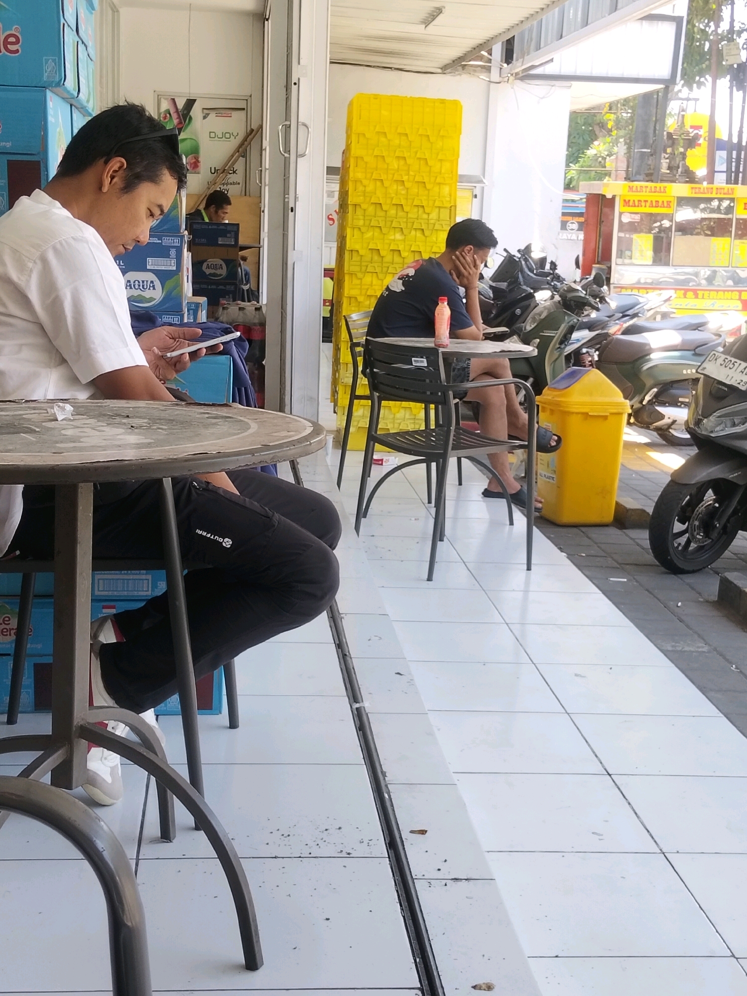 The image depicts an outdoor seating area in Kuta, Indonesia, likely at a small storefront or cafe. In the foreground, a man in a white shirt and black pants sits at a round, weathered metal table, holding a white smartphone in his hands, appearing to be looking at it. To his left, stacks of blue bottled water cases are visible. Behind him, another man in a dark t-shirt and shorts is seated at a black metal chair, with his head resting on his hand, appearing to be contemplating. A pink bottle is on the table in front of him. In the background, several parked motorcycles are visible, including a dark scooter in the right foreground. A bright yellow trash can stands between the two seated men. The ground is tiled with white squares. A building with yellow plastic crates is visible behind the man in the dark t-shirt. The scene is lit by sunlight, casting shadows.
