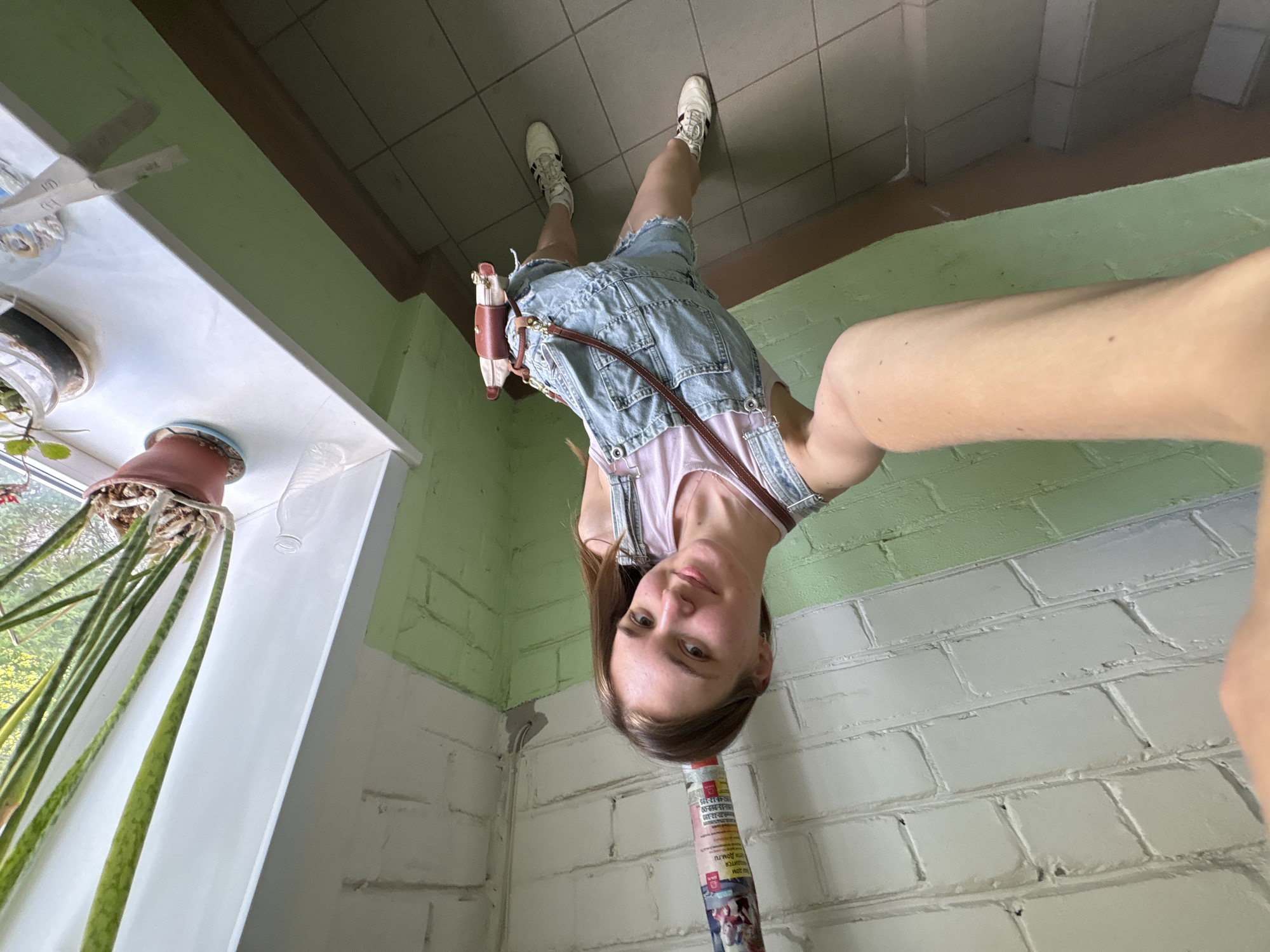A young woman with brown hair and light skin is taking a selfie in what appears to be an indoor space, possibly a residential building entrance or hallway. She is wearing light blue denim overalls over a light pink top and white sneakers with dark accents. She also has a brown purse with a tan strap. The woman is holding the camera at an angle, so the perspective is skewed.
The background includes walls painted with light green and white bricks. A pipe covered in what looks like a newspaper or advertisement is in the foreground near the bottom of the frame. To the left, there is a window with a white frame and a windowsill with a plant in a pot. The floor is covered with gray tiles.
Without specific visual cues or location data within the image, pinpointing the exact location in Perm, Russia, is impossible.