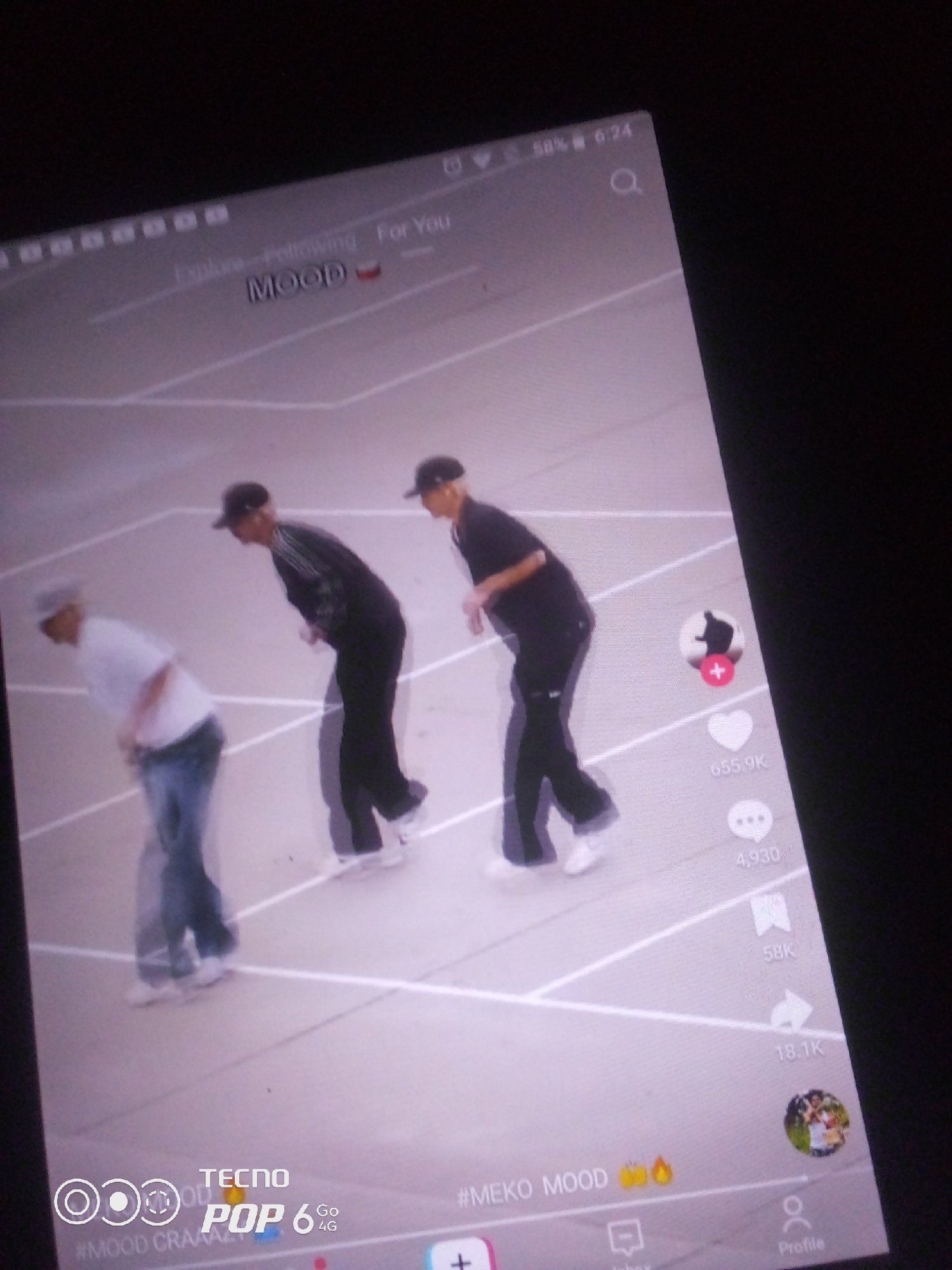 The image is a screenshot of a mobile device displaying a social media video. The video features three individuals dancing on an outdoor court with white lines. The first person on the left wears a white t-shirt and blue jeans. The second person in the middle wears a black tracksuit and a black cap, bending forward. The third person on the right wears a black shirt and a black cap, also bending forward. On the right side of the screen are social media engagement icons, including a heart with "655.9K" and a speech bubble with "4,930". At the bottom of the screen, text reads "#MEKO MOOD 🔥". The device is a TECNO POP 6 4G. The image is taken at an angle, suggesting the device is being held or placed at an incline. There is no information in the image to indicate the location in the city of Howayi, Nigeria.