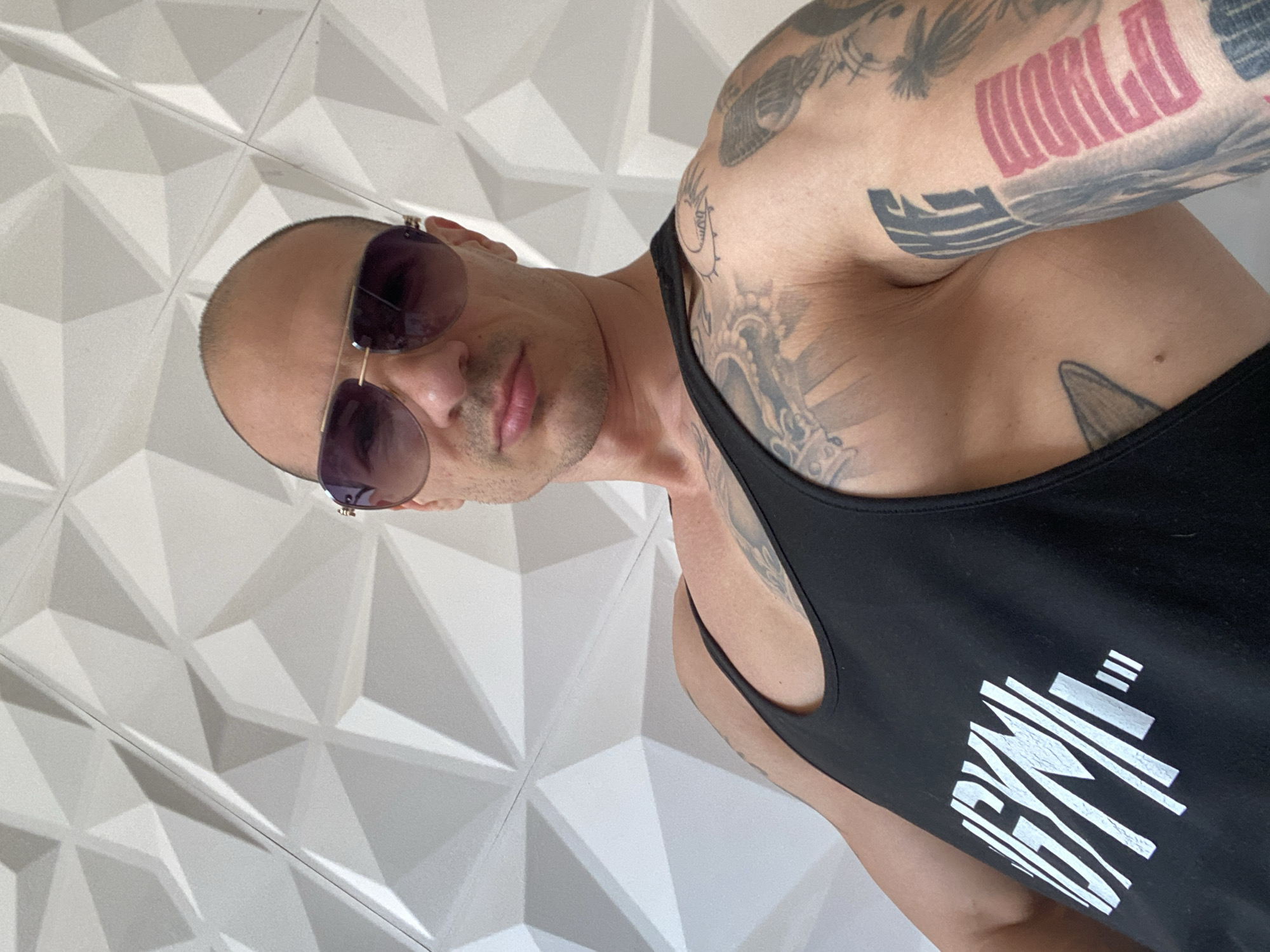 A bald man wearing sunglasses and a black tank top stands against a white, geometrically patterned wall. The man has numerous tattoos on his right arm and shoulder, including the word "WORLD" in pink block letters. He is positioned at an angle, facing towards the upper left corner of the image. The tank top has a white graphic design on the lower right side. The location appears to be indoors. The image does not provide information regarding Pleven, Bulgaria.