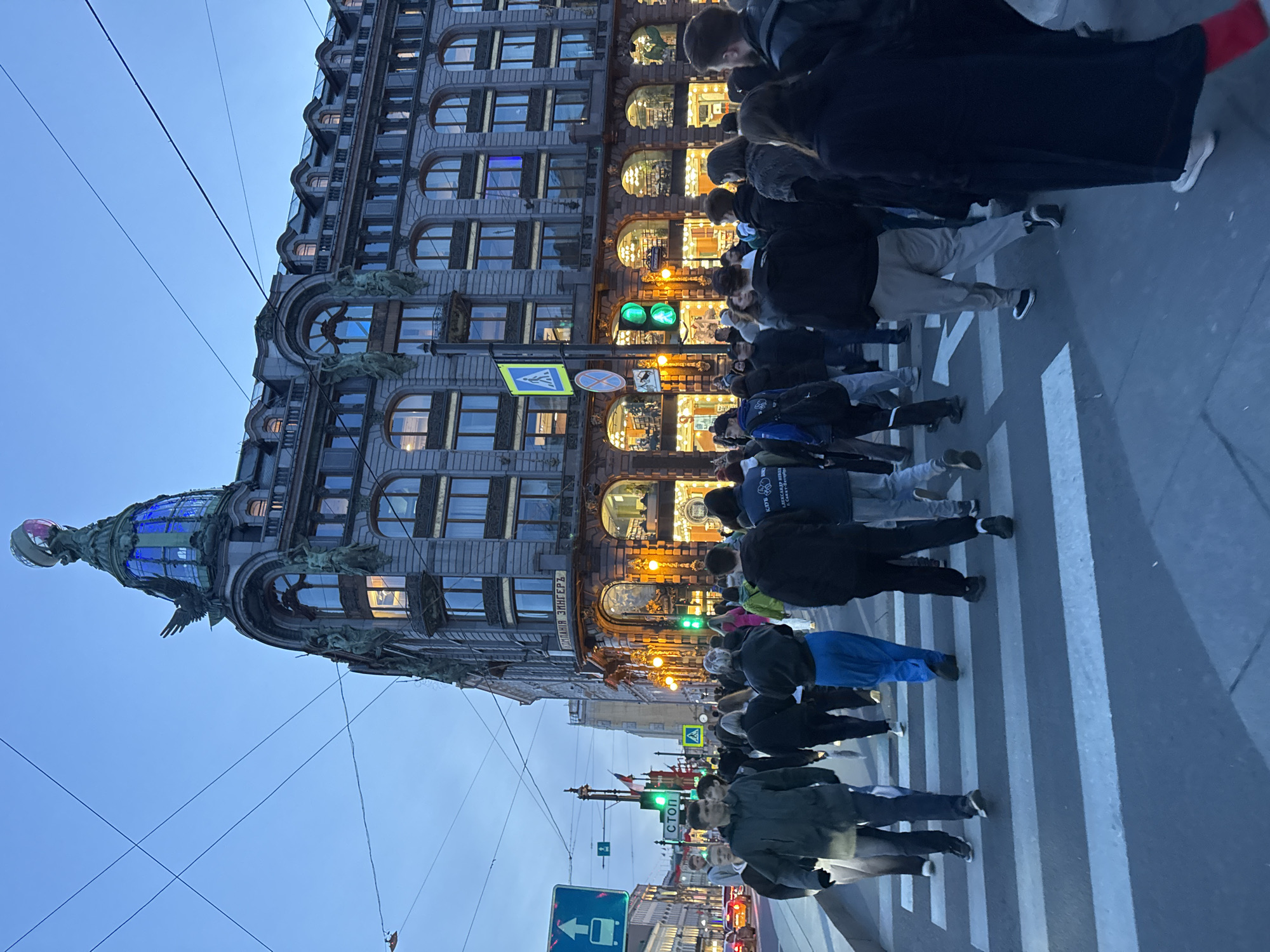 The image depicts a street scene in Saint Petersburg, Russia, featuring the Singer House, a prominent Art Nouveau building. A crowd of people is crossing the street at a pedestrian crossing. The traffic light is green, indicating it is safe for pedestrians to cross. The building is brightly illuminated, contrasting with the dusk sky. The architecture of the Singer House is notable, with a glass dome and ornate details. The sky is a light blue color, suggesting twilight. Utility wires are visible against the sky. Street signs and traffic signals are visible. The scene captures a typical urban environment.