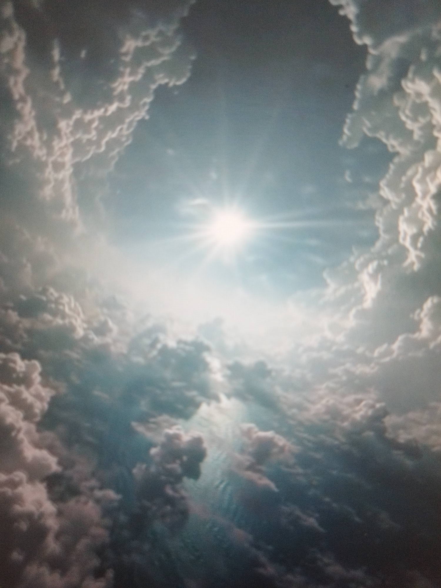 The image shows a view looking up into a sky filled with clouds. The sun is visible in the center, shining brightly. The clouds are a mix of light and dark shades, creating a textured and somewhat dramatic scene. There is no evidence to suggest that this picture was taken in Sofia, Bulgaria.