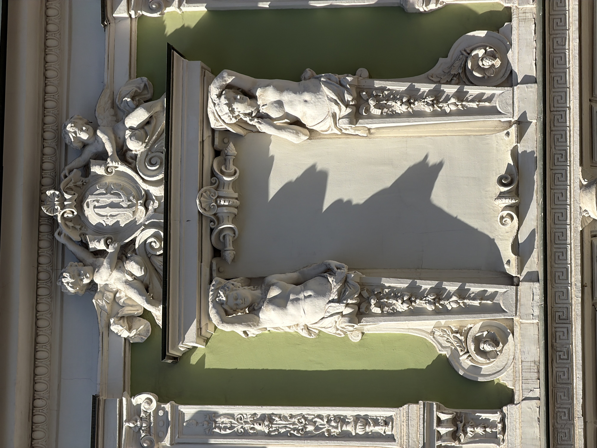 This is a close-up shot of a building facade, likely in Moscow, Russia, featuring intricate architectural details. The dominant elements include sculpted figures of reclining females on either side of a blank panel, flanked by ornate decorative moldings and relief carvings. On the left side, there is a sculpture of two figures, possibly cherubs or angels, holding what appears to be a shield or emblem. The facade is a light color, possibly white or off-white, and is accented by a light green paint. The lighting suggests a sunny day, with strong shadows cast across the facade, highlighting the three-dimensional nature of the sculpted elements. The carvings and sculptures appear to be of classical or neoclassical style.