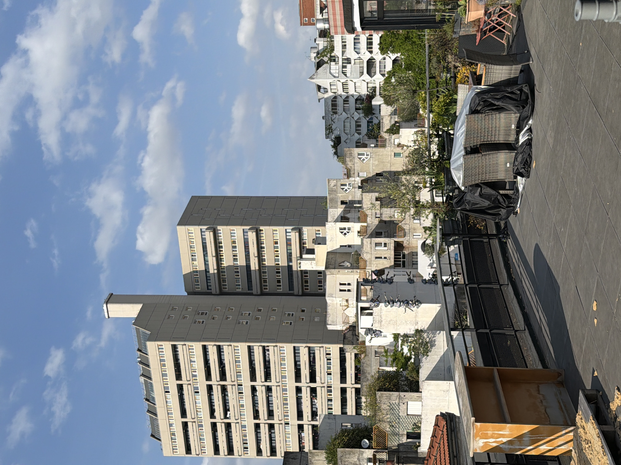 The image captures a rooftop scene in Ivry-sur-Seine, France, juxtaposing domestic elements with urban architecture. The foreground is dominated by a dark, flat rooftop surface, on which outdoor furniture, partly covered with black tarps and a silver tarp, sits. There are other objects, including what appears to be some sort of storage container, likely for use on the roof.
In the midground, a cluster of residential buildings and high-rise apartments form the city backdrop. One distinct architectural feature is a modern building with a unique, geometric facade.
The sky, taking up a substantial portion of the frame, shows a partly cloudy sky. The lighting is bright and suggests a sunny day. The composition is oriented in an unusual portrait shot, adding an artistic element to the scene.