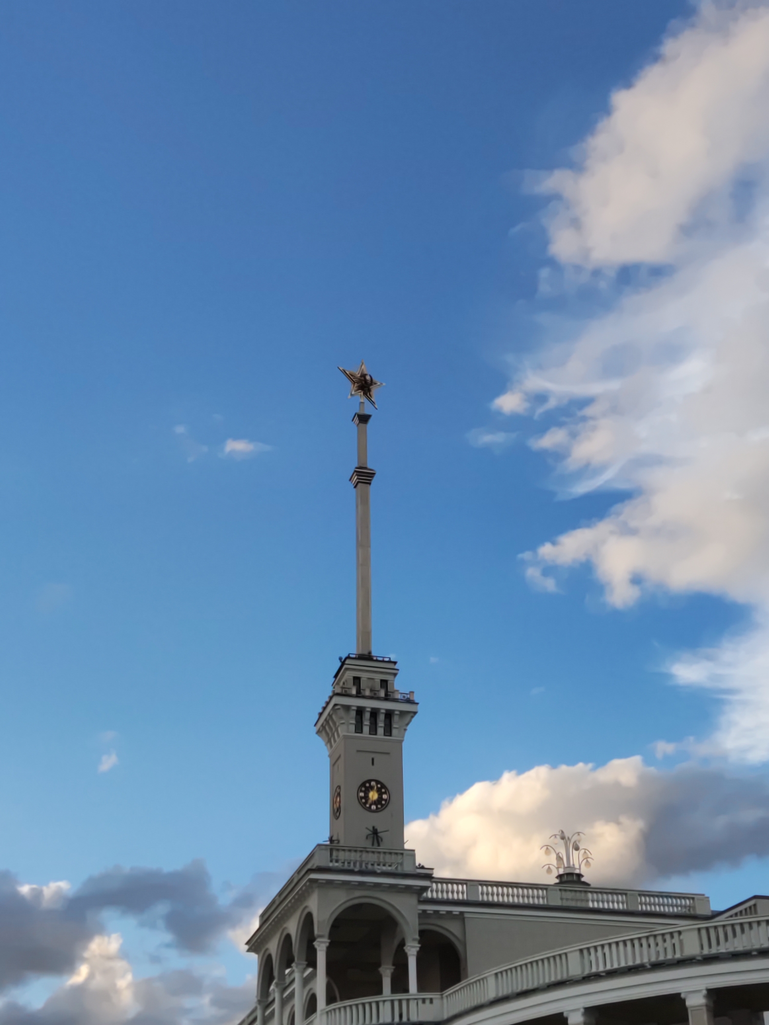 The image shows the North River Terminal in Khimki, Russia, against a backdrop of a bright blue sky with scattered clouds. A tall, slender tower with a clock face and a star ornament at its pinnacle dominates the center of the frame. The lower portion of the building features arched colonnades and balconies with white railings. A decorative lamp post with multiple lights is visible on the rooftop to the right. The lighting suggests it is daytime, possibly late afternoon.