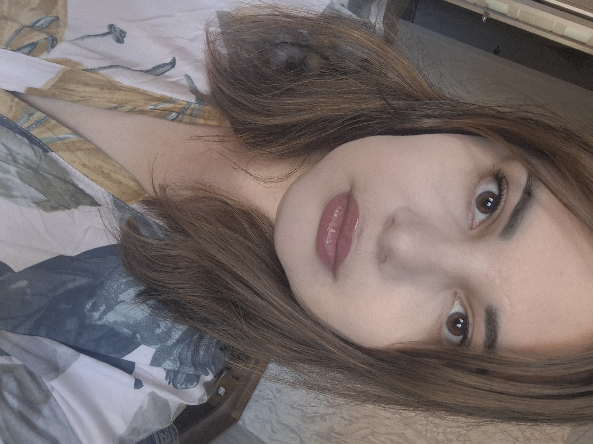 The image is a close-up selfie of a person with light skin, brown hair, and brown eyes. The person is lying on a patterned bedspread with floral and abstract designs in shades of white, blue, green, and yellow. The background shows a glimpse of a light-colored wall and a dark object that appears to be furniture. There is no information available in the image to suggest a connection to Амзя, Russia.