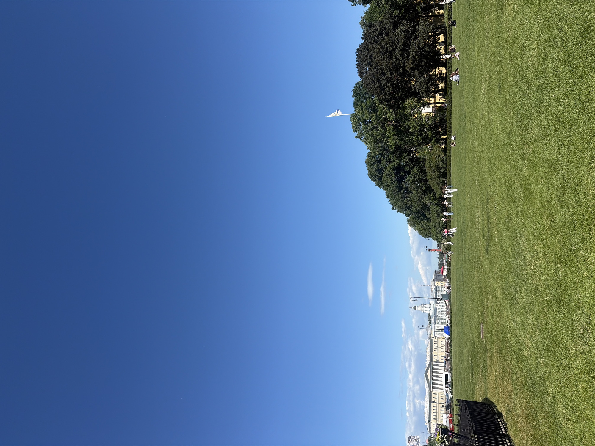 The image depicts a wide, manicured green lawn in the city of Saint Petersburg, Russia, under a clear blue sky with sparse, wispy clouds. In the background, a line of imposing classical buildings with colonnades stretches across the horizon. To the right of the buildings, a dense cluster of mature trees, some dark green and others with a slightly reddish hue, forms a natural backdrop. A white flag with a blue and yellow emblem flies atop the trees on the right side of the frame.
Several individuals are scattered across the lawn. Some are standing and appearing to engage in casual conversation or observe their surroundings, while others are seated or lying down on the grass, suggesting leisure activities like picnicking or sunbathing. A black metal fence runs along the left edge of the frame, bordering the lawn. Lampposts are visible along the path in front of the buildings. The scene conveys a sense of a peaceful, public park or square within the historic urban landscape of Saint Petersburg.