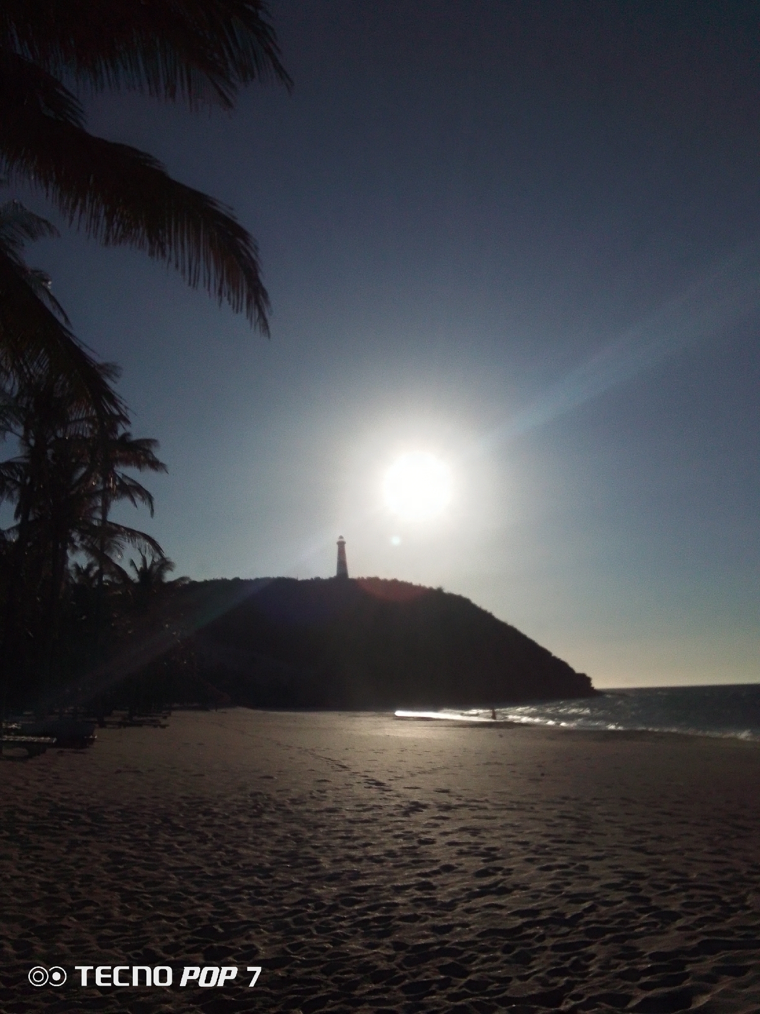 The image depicts a beach scene in Pedro González, Venezuela, at what appears to be dusk or dawn. The sun is directly overhead, emitting strong rays, and partially obscuring a lighthouse located atop a dark, elevated landmass. Palm trees line the beach on the left side. The beach itself is dark and sandy, with visible patterns. Waves are breaking along the shoreline. The image is taken with a TECNO POP 7 smartphone.