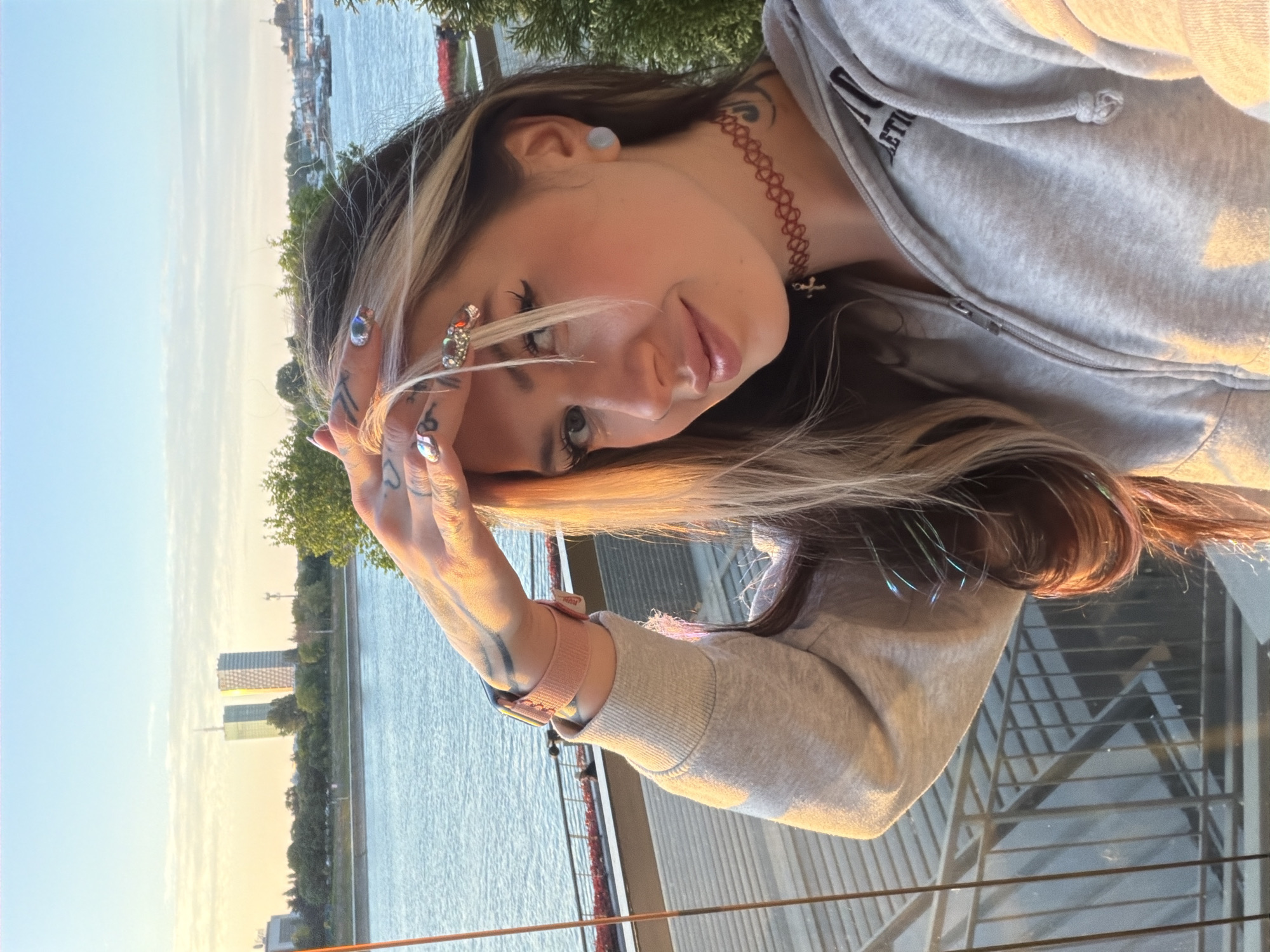 A woman poses for a selfie on a balcony overlooking the Sava River in Belgrade, Serbia. In the background, modern buildings line the riverbank. The woman is wearing a grey hooded sweatshirt and has a choker necklace and tattoos on her hand and neck. She is holding her hand up to shield her eyes from the sun. Her nails are decorated with rhinestones.