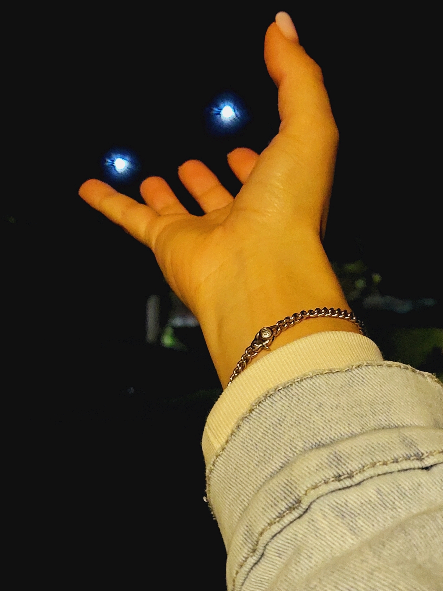 A hand is held up in the dark, with two small blue lights hovering above it. The hand is illuminated from the side, casting a warm glow. A silver bracelet with a delicate chain is worn on the wrist, over a light-colored garment, likely a long-sleeved shirt. The background is dark and out of focus, suggesting a nighttime setting. The scene could be interpreted as a moment of wonder or magic. The location is Sozopol, Bulgaria, though the specific setting within the city is not identifiable.
