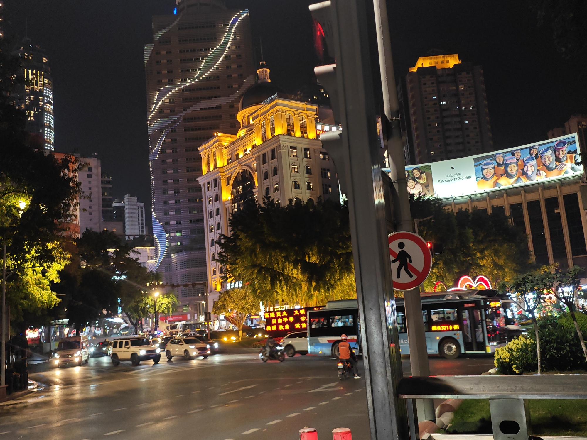 The image captures a street scene at night in what appears to be the Yuzhong District of China, judging by the architectural style and street signage. The photograph is taken from a lower angle, possibly at street level. In the foreground, a partially obscured "no pedestrian" sign is visible. Several vehicles, including SUVs, cars, and buses, are present on the wet, reflective street, suggesting recent rain. Pedestrians are also present, including a person on a motorbike wearing an orange shirt. Streetlights and building illuminations cast bright yellow glows, reflecting off the wet road surface. Tall, modern buildings dominate the backdrop, with a particularly prominent one featuring a wavy pattern of lights along its facade. A billboard displays a group photo and text in Chinese, potentially advertising the iPhone 17 Pro. The scene indicates a busy urban environment at night, with various forms of transportation and commercial activity.