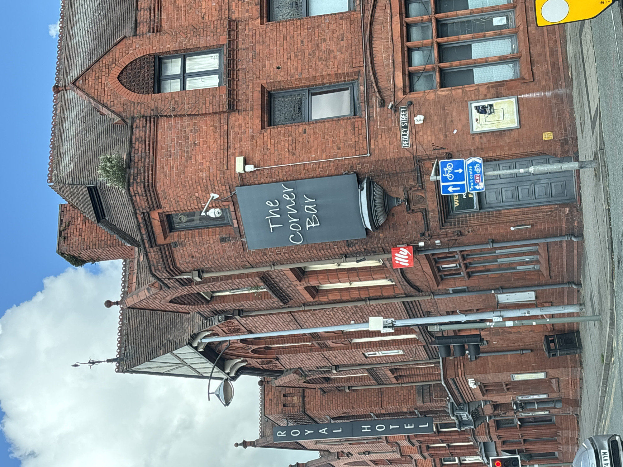 The photograph captures a street scene in Crewe, United Kingdom, featuring brick buildings. At the bottom, the words "ROYAL HOTEL" are displayed. Ascending upwards, there is a set of traffic lights and street paraphernalia such as lampposts. Above this, "The Corner Bar" has a sign, and further up is an "illy" sign. On the right, "PEDLEY STREET" is marked on a sign, along with cycling directions. The sky is partially visible.