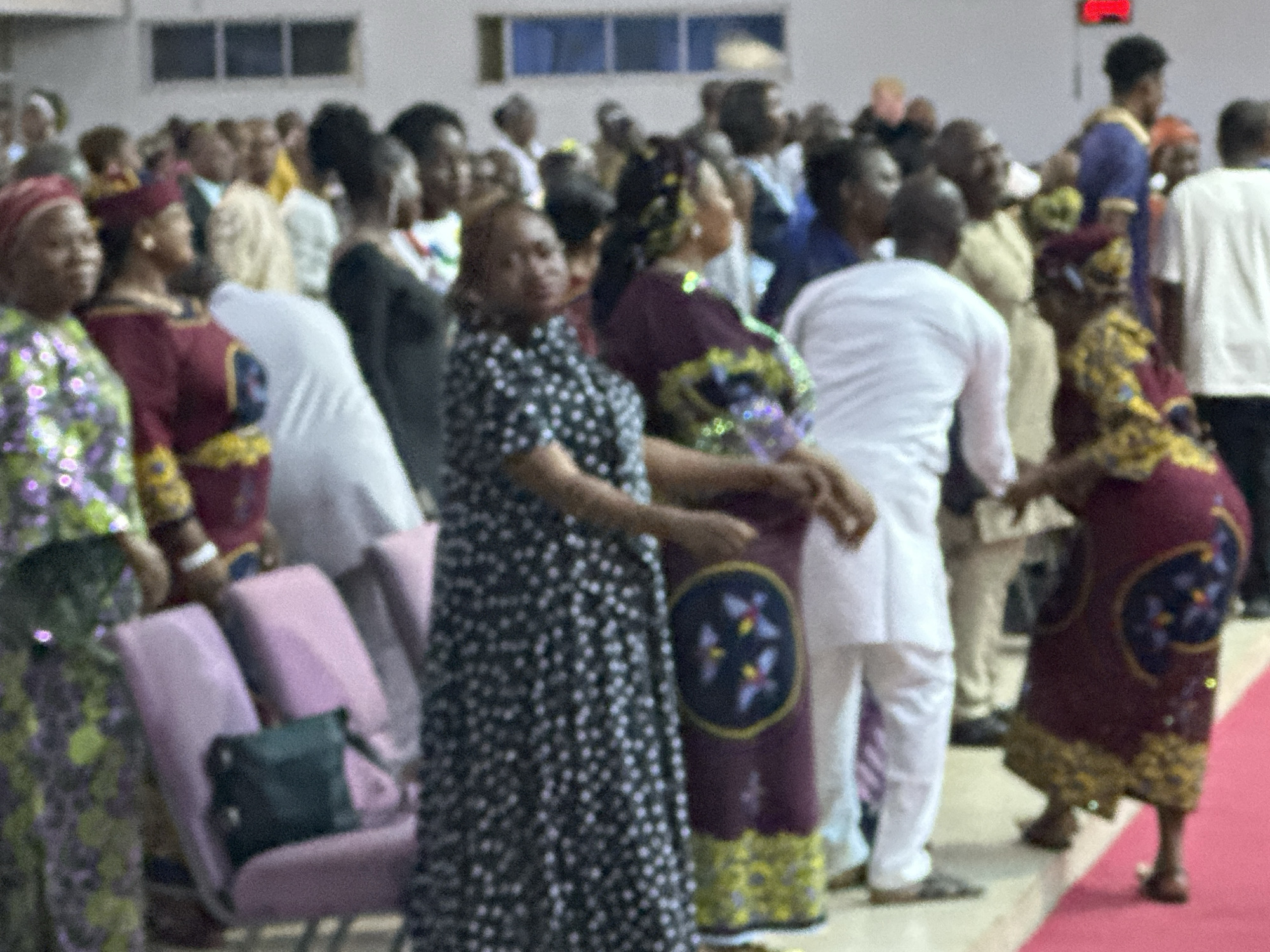 In Television, Nigeria, a crowd of people are gathered indoors, likely for an event or service. The image, taken from a slightly elevated perspective, focuses on the middle ground where several individuals are visible.
In the foreground, a woman wearing a black and white polka dot dress stands on the left, with her arms positioned downwards. To her right, another woman in a maroon and yellow patterned outfit is partially visible, also with her arms down. The seating area in front of them features several pink chairs with a dark green purse placed on one.
The middle ground of the image showcases a diverse group of people. On the far right, a woman in a maroon dress with elaborate gold and blue patterns is standing, her feet visible. She is facing towards the left and her hands are positioned in front of her. Next to her, a person in a white robe and pants is standing and facing away from the camera. To the left of this person, a woman in a dark, patterned dress is also visible. Further back, a man in a light yellow garment and a man in a dark blue robe are standing.
The background is blurred, showing more individuals in the distance. The environment appears to be a large hall with a light-colored wall and a few rectangular windows above. The floor seems to be a combination of a light surface and a reddish carpeted area towards the right. A red digital display is visible on the back wall. The overall impression is one of a gathering or a religious service in a community hall.