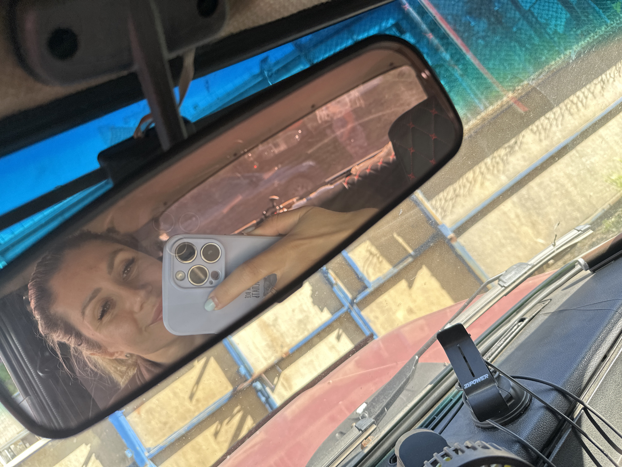 The image shows a selfie taken by a woman in a car, reflected in the rearview mirror. She is holding a light blue smartphone with a camera bump of four lenses in her right hand. She has blonde hair pulled back and is wearing makeup. The background outside the car appears to be a sunny day with a light-colored surface, possibly a parking lot or road. The interior of the car is visible, including the dashboard and a car mount with the logo "ZIPOWER". The image does not provide any specific details that would allow for the precise location within the city of Novoaltaysk, Russia.