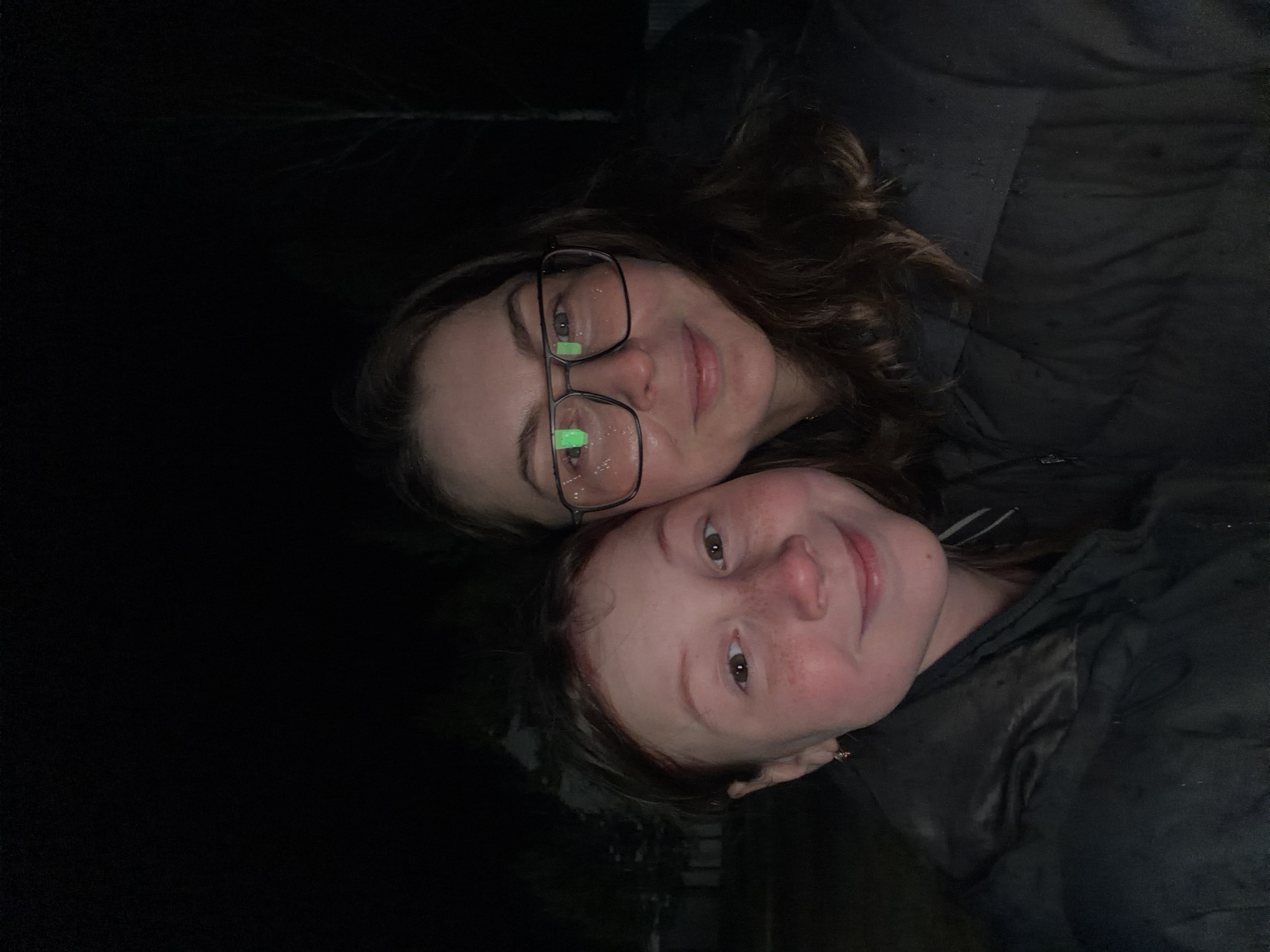 The image is a close-up portrait of two individuals, likely a mother and child or two women, taken outdoors at night in Markovo, Russia. The subjects' faces are illuminated, contrasting sharply with the extremely dark background.
The person positioned higher in the frame has dark, wavy hair and wears black-rimmed glasses. Green light reflections, possibly from a phone screen or LED source, are visible in her lenses. She has a subtle, gentle smile. Her head is leaning against the other individual's head or shoulder.
The second individual, positioned lower and to the right, has auburn hair and numerous visible freckles across her nose and cheeks. She also sports a warm, open smile, looking directly towards the camera. Both subjects appear to be wearing dark, possibly black, outerwear or jackets, suggesting cool or cold weather conditions.
The background is almost entirely black, with only faint, indistinct outlines of tree branches or sparse foliage barely discernible in the upper left corner. This indicates an outdoor, nighttime setting. There is no visible text in the image. The overall mood conveyed is one of affection, closeness, and contentment.