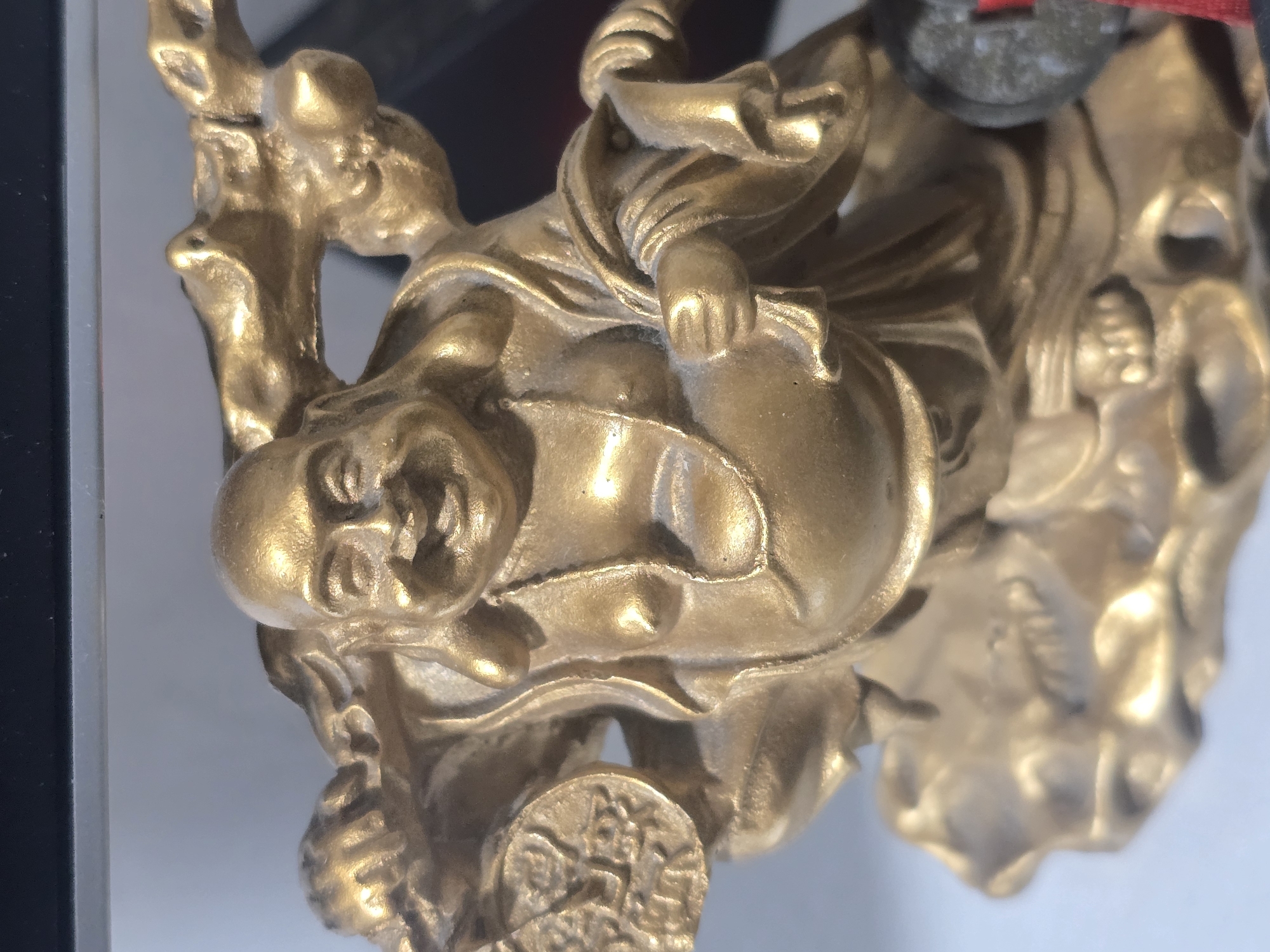 A close-up, eye-level, medium shot captures a golden figurine of a laughing Buddha, likely a decorative item. The figurine is made of a metallic material with a polished sheen, reflecting light. The Buddha is depicted with a round, smiling face, a prominent belly, and is draped in flowing robes. He is shown in a kneeling or squatting pose, holding a circular object with intricate carvings in his left hand. The figurine is placed on an irregular, textured base that resembles a natural rock formation. The background is softly blurred, showing a light-colored surface and darker, indistinct shapes, suggesting an indoor setting, possibly a shelf or display. There are no people present in the image. The setting is not identifiable as Ulan-Ude, Russia, or any specific city.