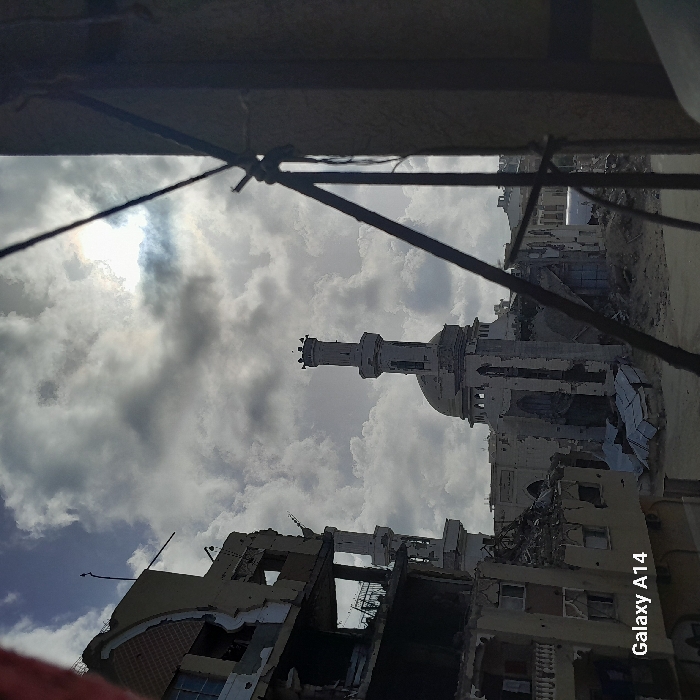 A devastated cityscape reveals a mosque's minaret amidst extensive building ruins. Structures show severe damage, potentially from conflict or disaster. The sky is overcast with scattered clouds. Wires and framing in the foreground suggest the perspective is from another damaged building. The overall scene conveys destruction and loss.