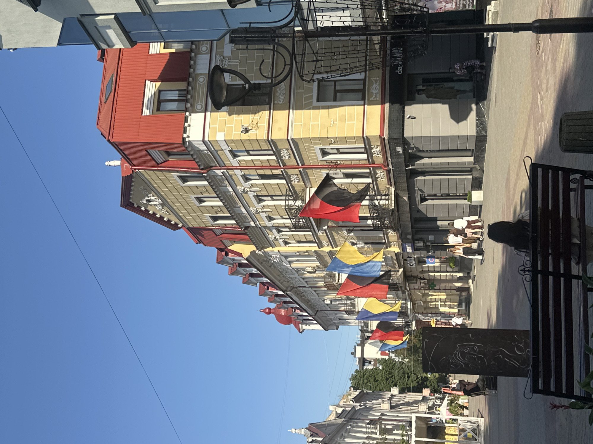 This media file depicts a street scene in Ternopil, Ukraine. The orientation of the image is rotated 90 degrees counter-clockwise, so the scene is viewed from a sideways perspective. The foreground features a black metal bench and a dark, ornate panel with intricate carvings of a face.
The background shows a street lined with buildings featuring colorful facades and architectural details. A string of flags in various colors, including black, red, yellow, and blue, are hung across the street, fluttering in the breeze. These flags may represent different nationalities or regions.
Several people are visible on the street. Some are standing near shop entrances, while others appear to be walking or sitting. One person is sitting on a bench in the foreground, partially obscured by shadows. Another person in dark clothing is visible in the middle ground, near a shop entrance.
The time of day appears to be daytime, with clear blue skies and bright sunlight casting shadows. The weather seems pleasant.
Visible text includes "CO.UA" on a sign in the background. The overall impression is that of a lively urban environment on a sunny day, likely a public space or pedestrian area within the city.