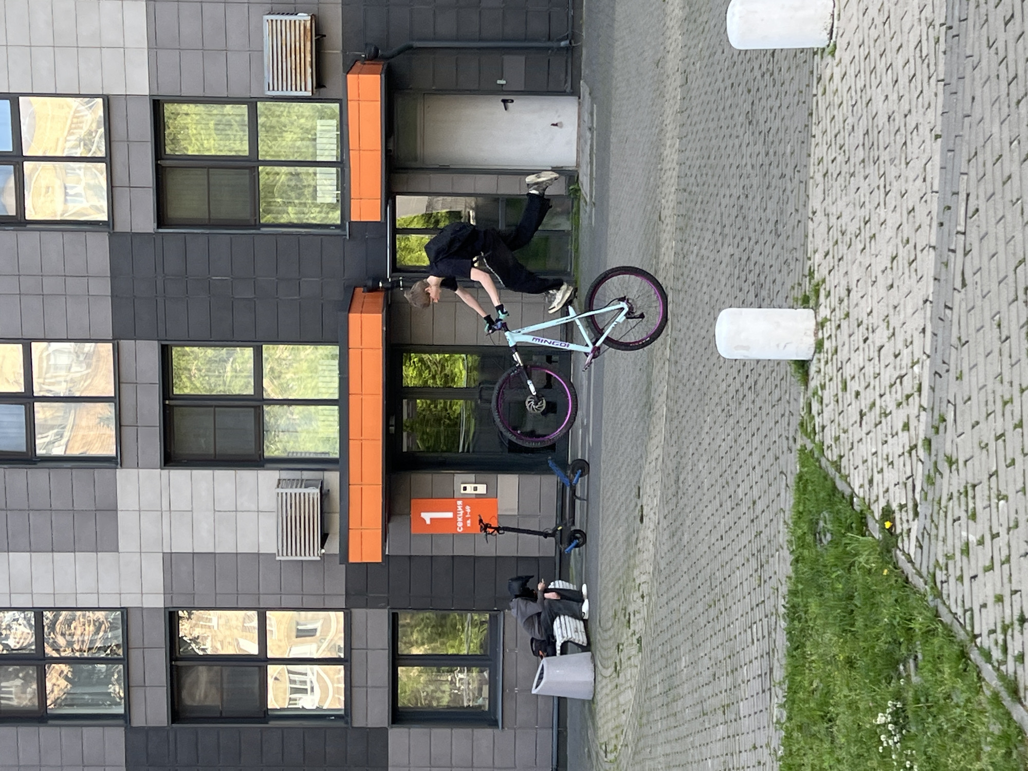 A young male is performing a stunt on a light blue bicycle with a purple rim, balancing on the rear wheel in front of a modern apartment building. He is wearing a black t-shirt, black pants, and a backpack. The building has a gray tiled facade with large windows reflecting trees and other buildings. Orange panels are visible on the building's exterior. To the left, a person wearing a helmet and dark clothing is sitting on a bench next to a trash can, looking at their phone. Two electric scooters are parked nearby. The scene takes place outdoors on a paved area with some grass patches and white bollards. The location is Mytishchi, Russia.