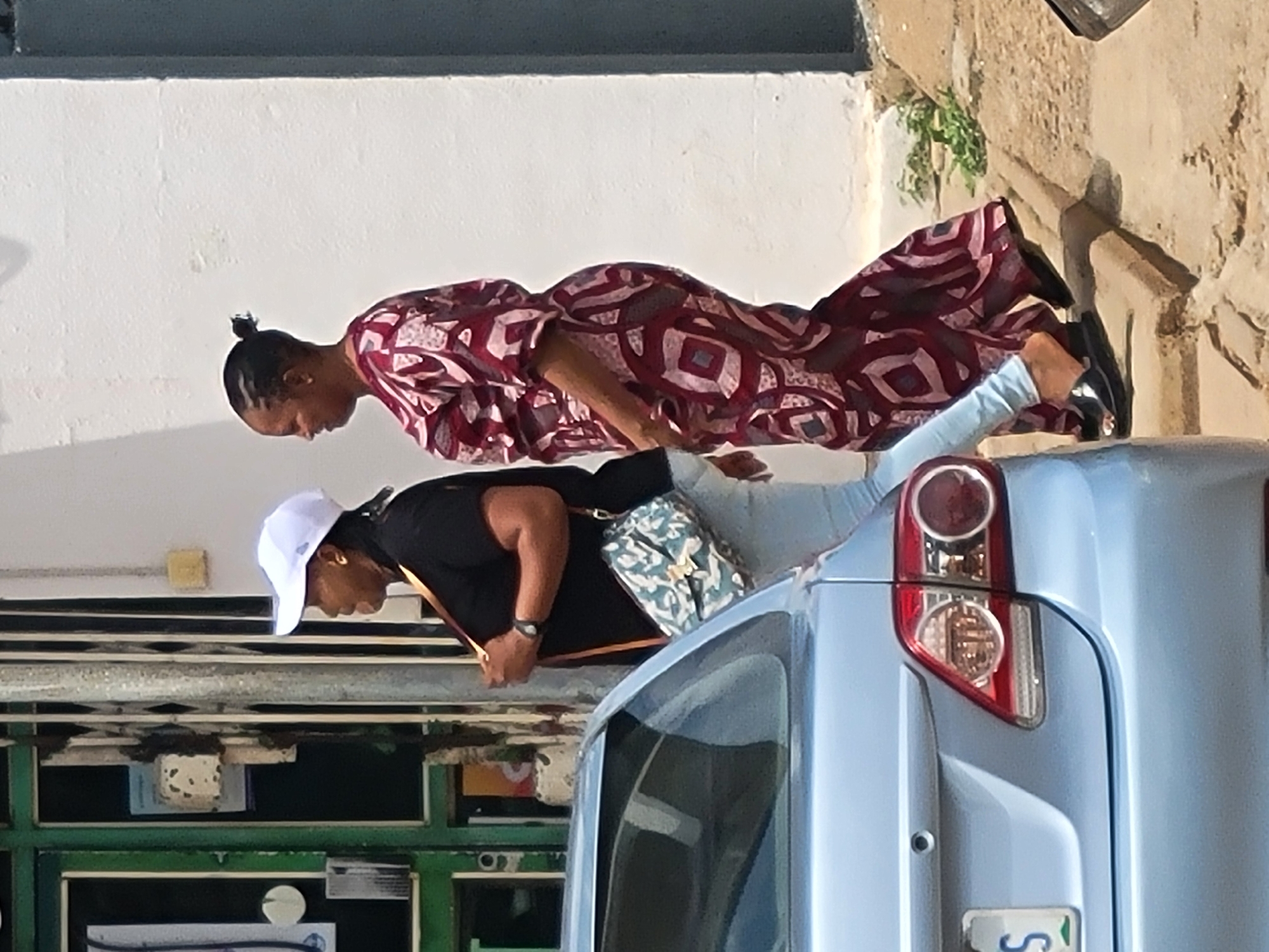 Two African women stand next to a light blue vehicle. The woman closest to the vehicle wears a black shirt, light blue jeans, a white baseball cap, and a crossbody bag. The second woman wears a maroon and white patterned dress, light blue jeans, and black shoes. The vehicle appears to be parked in a city. The green and white building facade and natural vegetation indicate it is Jos, Nigeria.