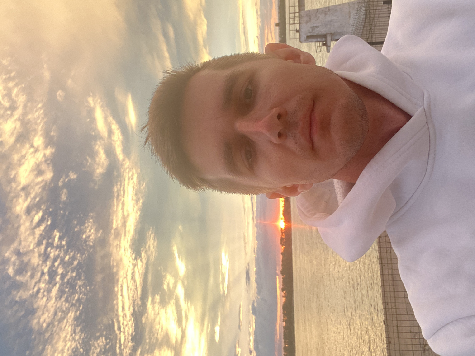 This is a selfie taken outdoors at dusk, likely in Novokuybyshevsk, Russia, as indicated by the presence of a body of water and an industrial structure. The foreground features a man from the chest up, wearing a white hooded sweatshirt. He has short, brown hair and is looking directly at the camera with a neutral expression. His face is well-lit by the ambient light.
In the background, a wide body of water stretches across the scene, reflecting the warm, golden hues of the setting sun. The sky is filled with dramatic clouds, tinged with orange and yellow, suggesting the sun is low on the horizon. A tree-lined shore is visible in the distance, and a small industrial structure with some scaffolding or a platform is situated near the right edge of the frame, close to the water's edge. The overall atmosphere is serene and picturesque.