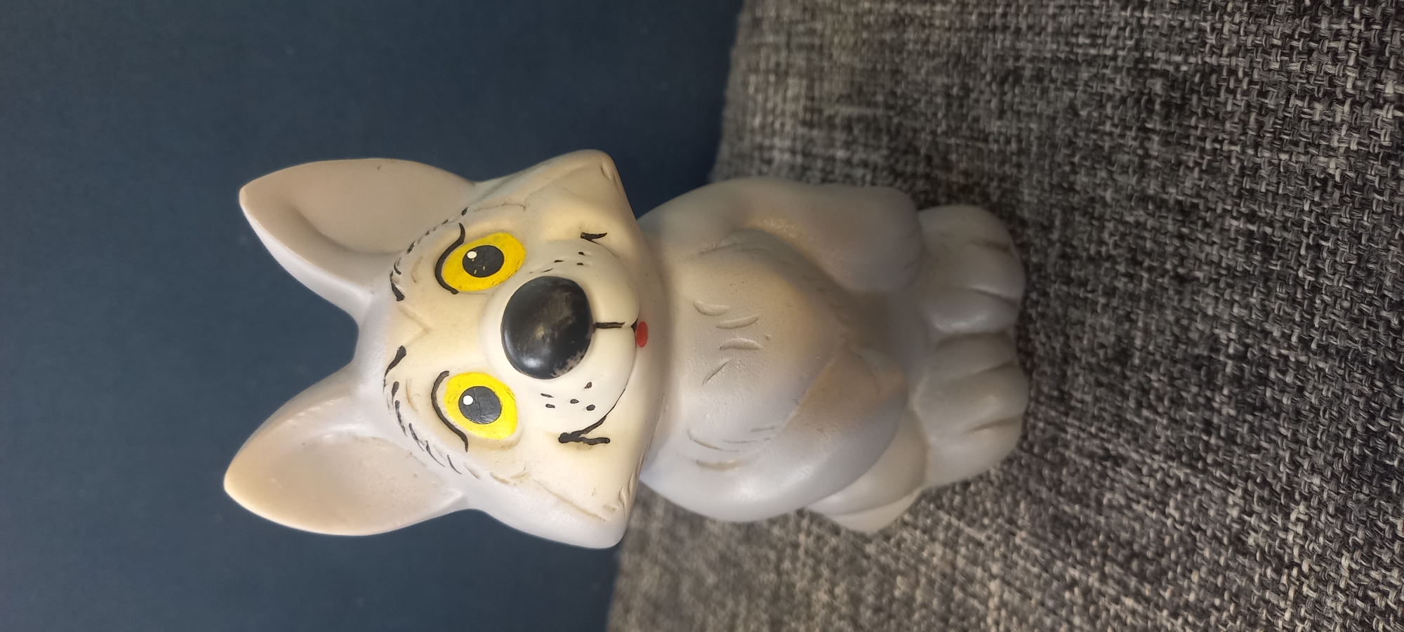 The image shows a small, light gray, plastic toy wolf figurine. The wolf has large, bright yellow eyes with black pupils, a black nose, and a small red mouth. It has black painted eyebrows and a few black dots painted on its cheeks, suggesting whiskers. The figurine is sitting on a textured gray fabric surface, which appears to be upholstery. Behind the figurine, a dark blue background is visible. The lighting suggests an indoor setting. There is no indication of the location in the city of Идрисово, Russia.