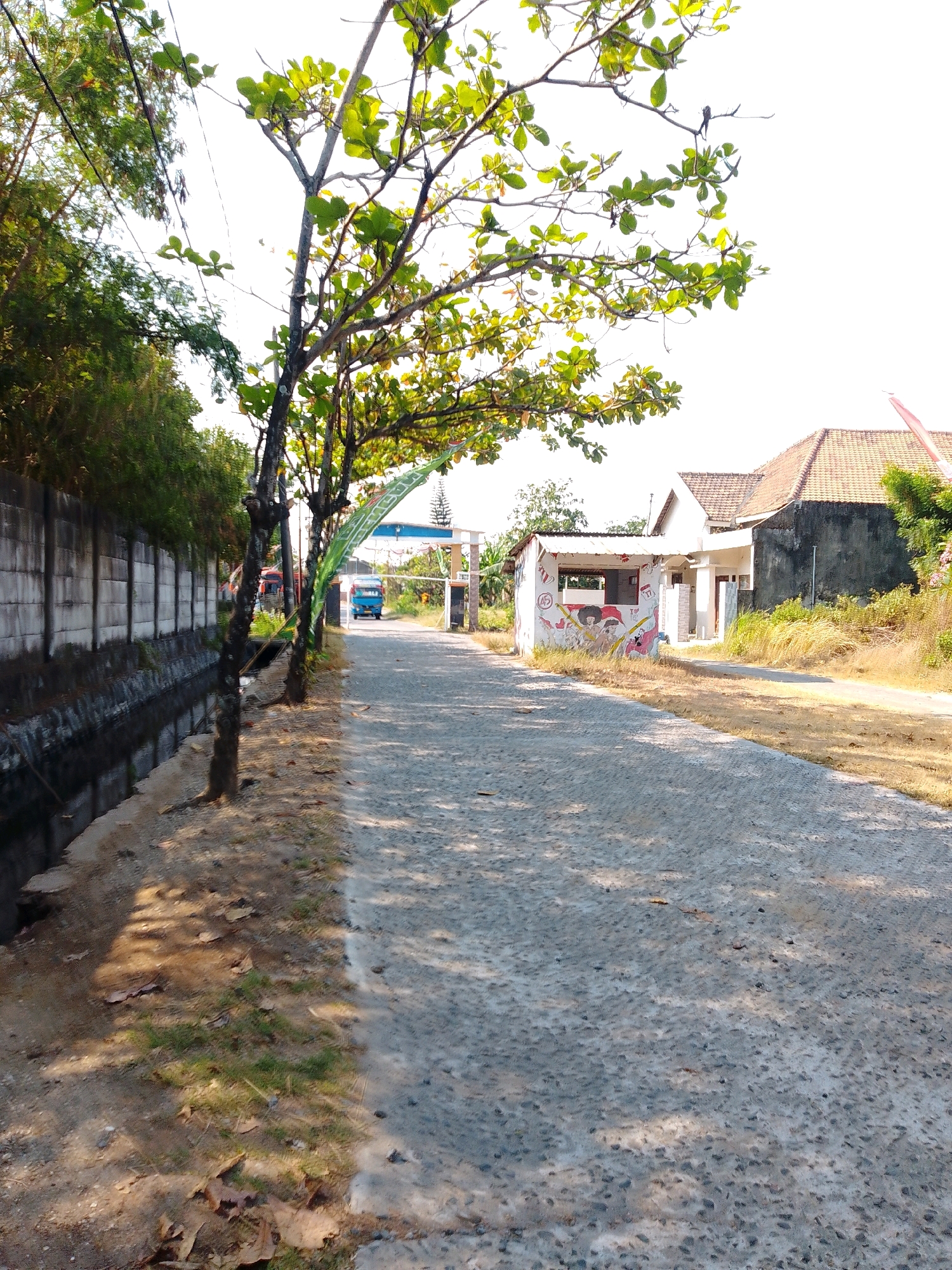 The image depicts a street scene in Cikal, Indonesia, on a sunny day. The road is paved with gray stones and is lined by a concrete wall on the left and trees on the right. A narrow channel of water runs alongside the wall. Further down the road, a small, white building with a red-and-white mural is visible. Beyond that, a blue truck is approaching a gateway. The sky is bright and overcast. The overall impression is of a rural or suburban area.