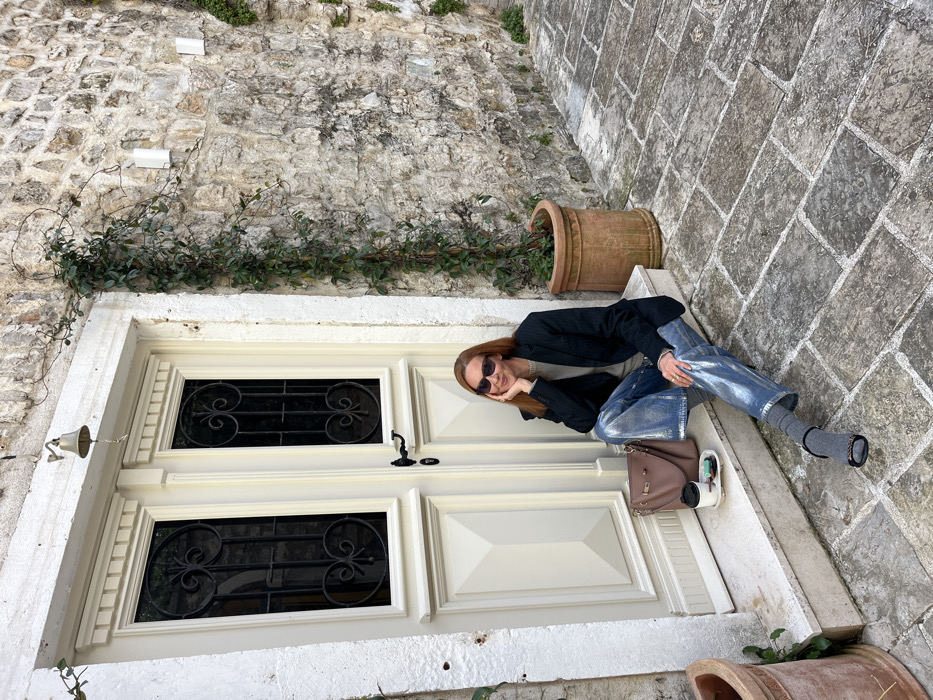 A woman sits on the lower stone step of a building entrance in Budva Old Town, Montenegro. She has reddish-brown hair and wears dark sunglasses, a light-colored top, a dark blazer, metallic-silver trousers, grey socks, and dark open-toed shoes. Her right hand is propping her chin, and her left arm rests on her lap. To her left, a taupe-colored bucket bag, a disposable coffee cup with a dark lid, and a small white dish containing a green object are placed on the step.
The background features a light-colored double-leaf wooden door with decorative molding and two glass panels fitted with dark, ornate metal grilles. A small brass bell hangs above the door. The building's exterior consists of an aged, rough-hewn stone block wall. A green climbing vine ascends the wall to the right of the door, alongside a terracotta pot containing plants. A second terracotta pot is partially visible on the far left. The ground is paved with irregularly shaped light grey cobblestones, forming a narrow street or alleyway.