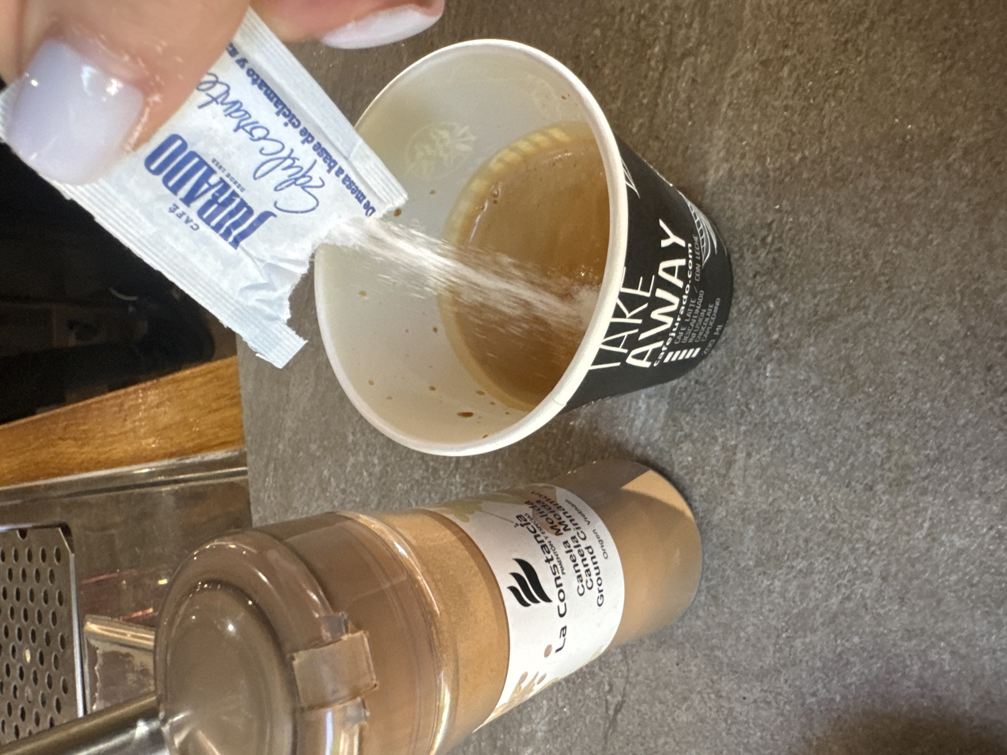 This is a close-up, top-down shot of someone preparing a coffee drink. The scene appears to be indoors, with a grey, textured countertop as the primary surface. To the left, a portion of a stainless steel coffee machine with a perforated drip tray is visible, along with a wooden surface.
The main focus is on a white paper cup with "TAKE AWAY" printed in black, containing a light brown coffee liquid. A person's hand, with a light purple-blue nail polish, is holding a small white packet labeled "JURADO Edulcorante" (sweetener) and is pouring its contents into the coffee. The packet also indicates "CAFÉ DESDE 1912" and some Spanish text about its composition. Beside the cup is a clear plastic bottle filled with a brown powder, identified by a label as "La Constancia Canela Molida Ground Cinnamon," originating from Vietnam.
The lighting suggests it could be daytime, possibly indoors in a cafe or kitchen setting. The overall impression is one of a personal moment of preparing a beverage, adding sweetener and cinnamon to a coffee. The visible text provides specific brand and product information, hinting at the location being Spain, given the Spanish language and specific product names.