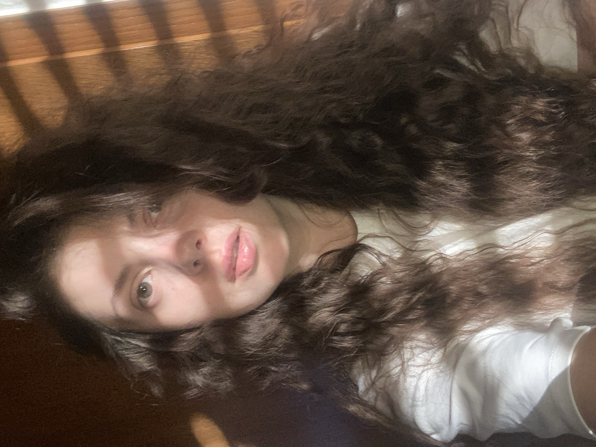 A woman with long, curly brown hair lies on a surface, possibly a bed or floor. She is wearing a white top. Light and shadows from what appears to be window blinds are cast across her face and the surrounding area. The setting's location in Landau in der Pfalz, Germany, is not evident from the image.