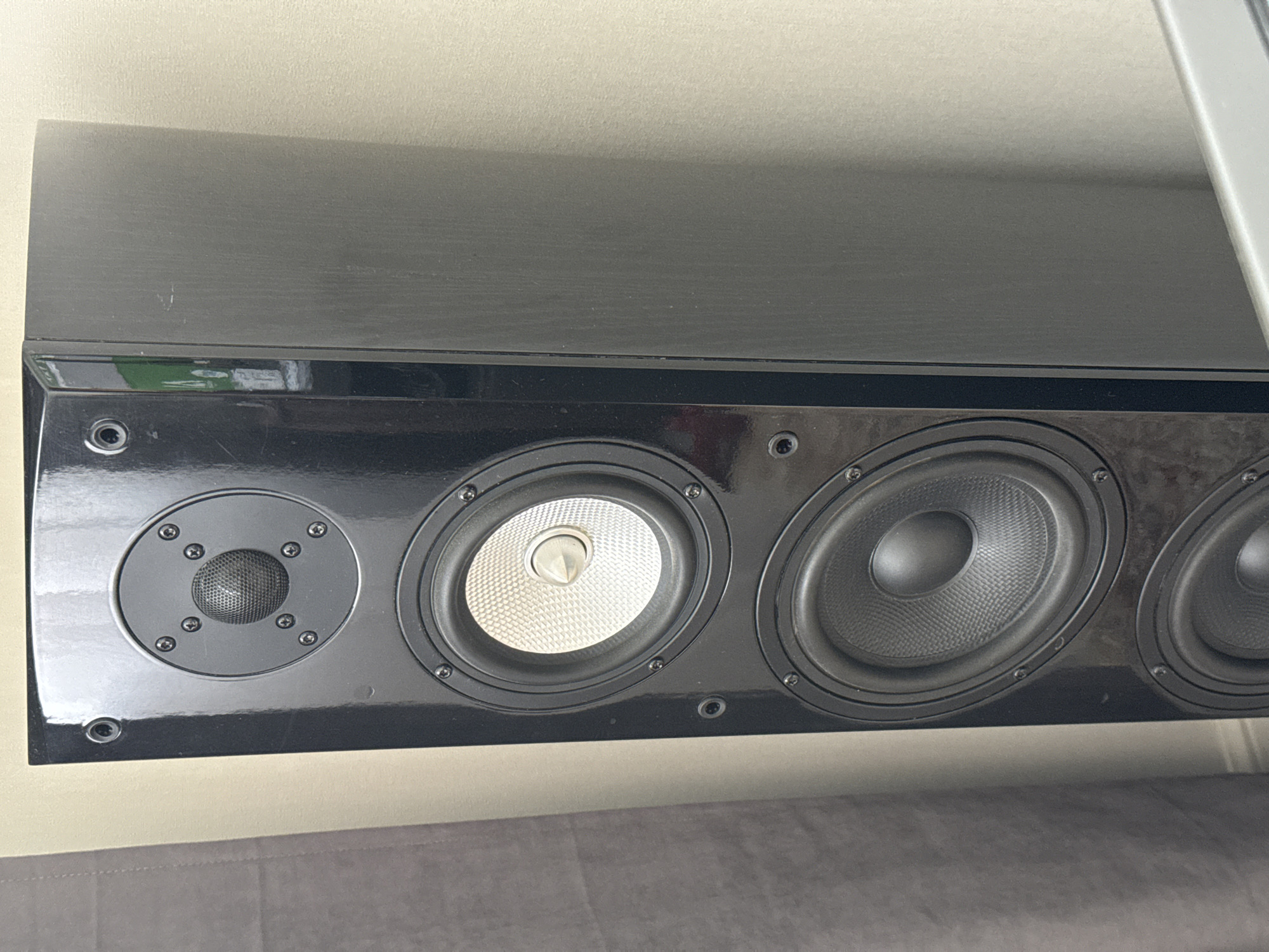 This is a close-up, eye-level photograph of a black, rectangular speaker unit mounted on a wall. The speaker features a glossy black finish and is divided into three distinct sections. On the far left is a small, circular tweeter with a mesh cover, secured by several screws. To its right are two larger, circular mid-range or woofer speakers. The left speaker cone has a silver, textured pattern with a central silver dome, while the right speaker cone is black and textured. The entire front of the speaker unit is framed by a glossy black bezel, with visible screws around each driver and what appear to be small indicator lights. The speaker is positioned above a grey upholstered surface, possibly a couch or bench, which occupies the bottom portion of the frame. The background wall is a light, neutral color. The lighting suggests it is daytime, with soft illumination and no harsh shadows, indicating an indoor setting. There is no text visible in the image.
