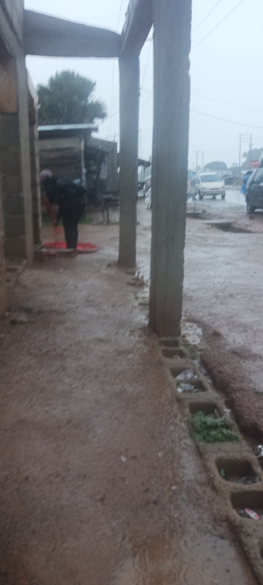 The image, taken in Akwanga, Nigeria, depicts a wet, possibly rainy, outdoor scene. In the foreground, there's a partially constructed or unfinished concrete structure with supporting pillars. A person is seen bending over, possibly working, near one of the pillars, with a red object lying on the ground nearby. Behind the person, there are some simple stalls or shops along the roadside. The road itself is wet and reflects the overcast sky. Cars are visible in the background, indicating vehicular traffic. A concrete drainage system runs along the edge of the walkway, filled with rainwater, debris, and some vegetation. The overall environment suggests a roadside market or commercial area during inclement weather.