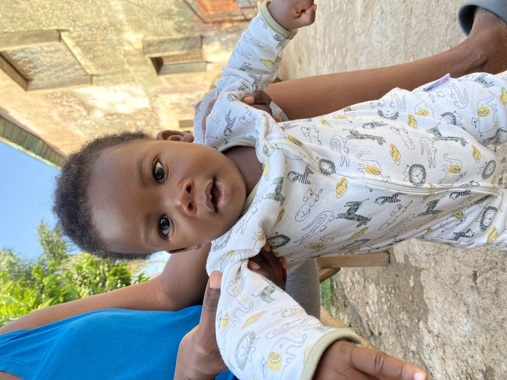 A close-up, eye-level, outdoor shot features a Black baby wearing a white onesie with a repeating pattern of cartoon animals, including elephants, lions, zebras, and crocodiles. The baby's face, framed by short, dark curly hair, is centered in the frame. Their eyes are open and looking forward, and their mouth is slightly ajar. The baby is being held by an unseen adult whose arms and torso, wearing a blue tank top, are visible on the left side of the image, and their legs, in dark pants, are on the right. The background depicts a weathered, light-colored building with visible windows and a green-trimmed roof. The ground is composed of dirt and loose stones. The scene is illuminated by natural sunlight, casting shadows on the ground and building. This image was captured in Sabon Tasha, Nigeria.
