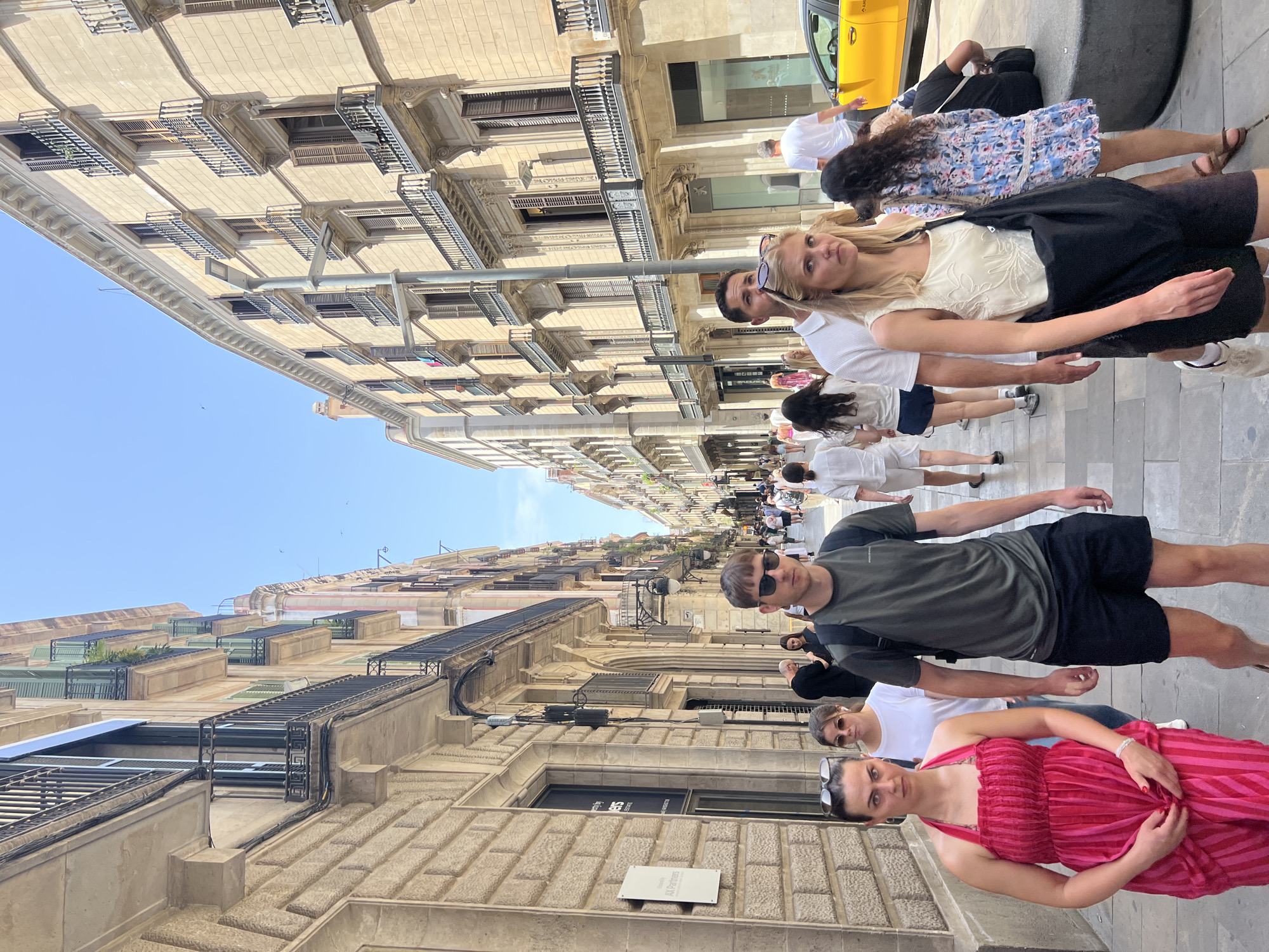 The image depicts a street scene in Barcelona, Spain, likely during the daytime given the bright sunlight. The perspective is at an oblique angle, with the camera pointing upwards towards the buildings and the sky.
Several people are visible, walking along the street. In the foreground, a woman in a vibrant pink and red striped dress is looking at the camera, her hand resting on her dress. Next to her, a man in a dark green t-shirt and black shorts is also looking towards the camera, wearing sunglasses. Further back, other individuals and groups are strolling along, some in casual attire. A yellow taxi is partially visible on the right side of the frame.
The architecture of the buildings flanking the street is a notable feature. They are tall, multi-story structures with ornate facades, featuring balconies with decorative railings. The buildings appear to be made of light-colored stone or stucco, and many windows are visible, some with shutters. The street itself is paved with cobblestones or similar material.
The overall impression is a bustling urban environment on a sunny day. The individuals in the foreground appear relaxed and aware of the camera, while the general atmosphere suggests a leisurely stroll through the city. There is no discernable text in the visible portions of the image.