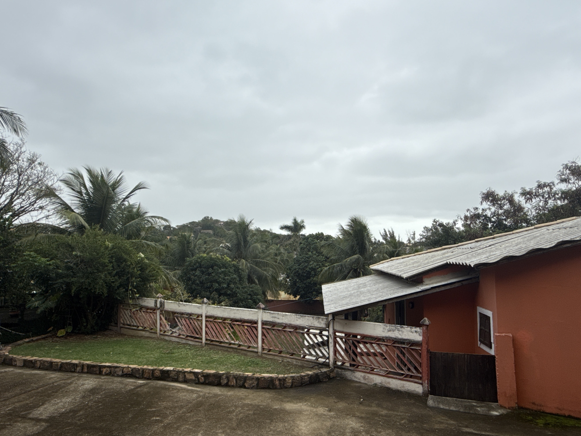 The image shows a view from an overcast day in São Pedro da Aldeia, Brazil. In the foreground, a dark, damp concrete area leads to a small grassy lawn bordered by a rough stone wall. A red and white metal fence with a diagonal lattice pattern separates the lawn from the background. Beyond the fence, lush green trees and palm trees dominate the landscape, with some visible on rolling hills in the distance. To the right, the corner of a bright orange building with a corrugated metal roof is prominent. The building has a small, dark-framed window. The sky is heavily clouded and gray.