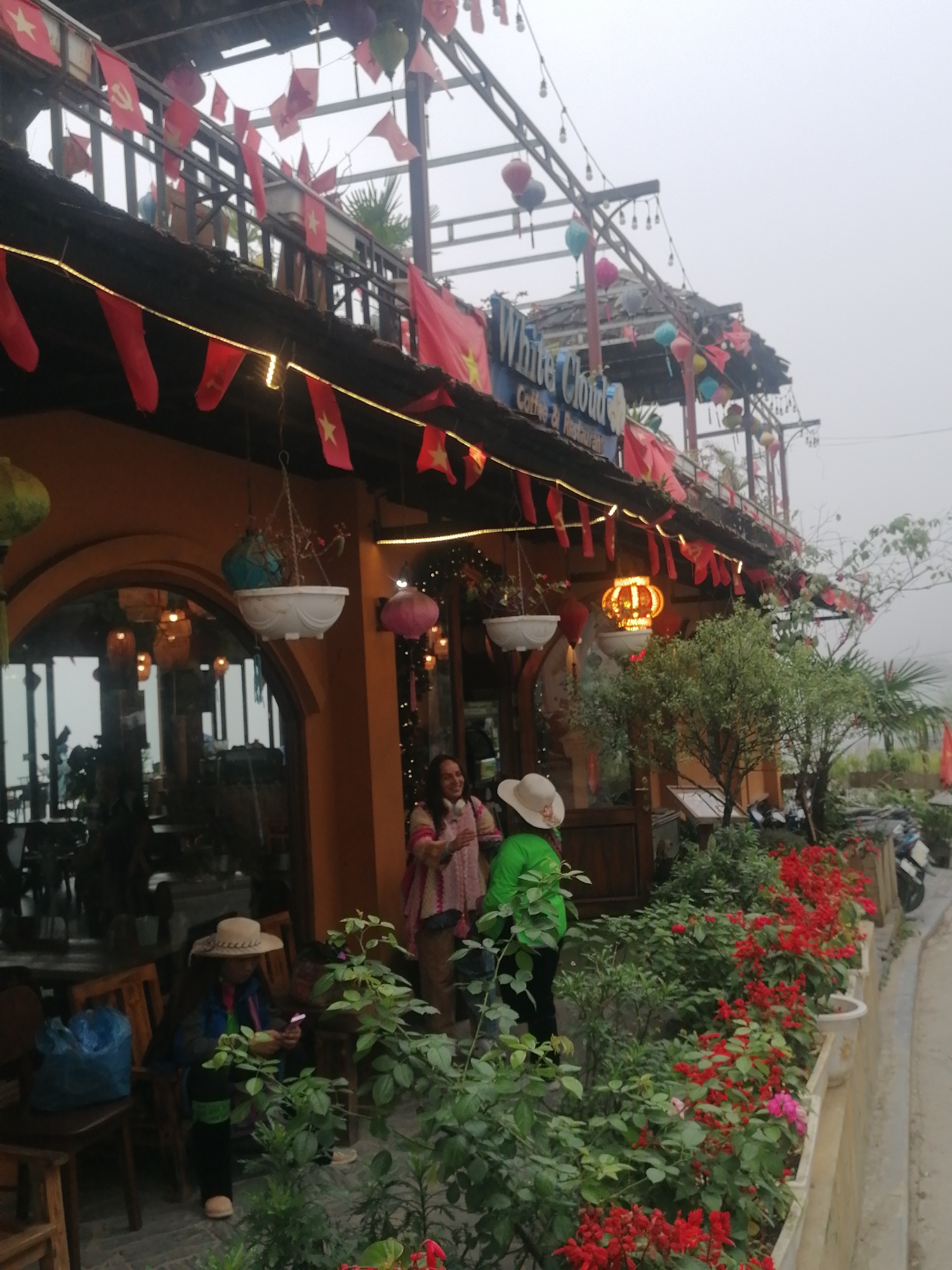 The image captures the exterior of "White Cloud Coffee & Restaurant" in Sa Pa Ward, Vietnam, on an overcast or foggy day. The two-story building has a warm, orange-toned facade on the ground floor, featuring arched windows and doorways, with an upper balcony adorned with decorative elements.
The cafe's name, "White Cloud Coffee & Restaurant," is visible on a blue sign above the entrance, illuminated by string lights. The establishment is heavily decorated with numerous small red flags. These include flags of Vietnam (red with a yellow star) and flags bearing the hammer and sickle symbol, indicating Communist affiliation. String lights are strung along the roofline, and colorful paper lanterns hang from the eaves and balcony structure. Hanging planters with greenery are placed near the entrance.
Three women are visible at the front of the cafe. One woman, wearing a straw hat and green top, stands near a garden bed. Another woman, also wearing a straw hat, sits at an outdoor wooden table, seemingly looking at a phone or small device. A third woman, wearing a light-colored patterned top and headphones, stands further back near the main entrance. The overall attire of the people suggests cool weather, consistent with the foggy conditions.
In front of the building, there are rows of vibrant red flowering plants in planters, creating a lush border. Other green potted plants and small trees are also part of the landscaping. Wooden tables and chairs are arranged for outdoor seating. In the background, beyond the plants and figures, the outlines of parked scooters or motorcycles can be faintly seen on the paved area next to the building. The sky is uniformly light gray, indicating thick cloud cover or fog, typical of mountainous regions like Sa Pa.
