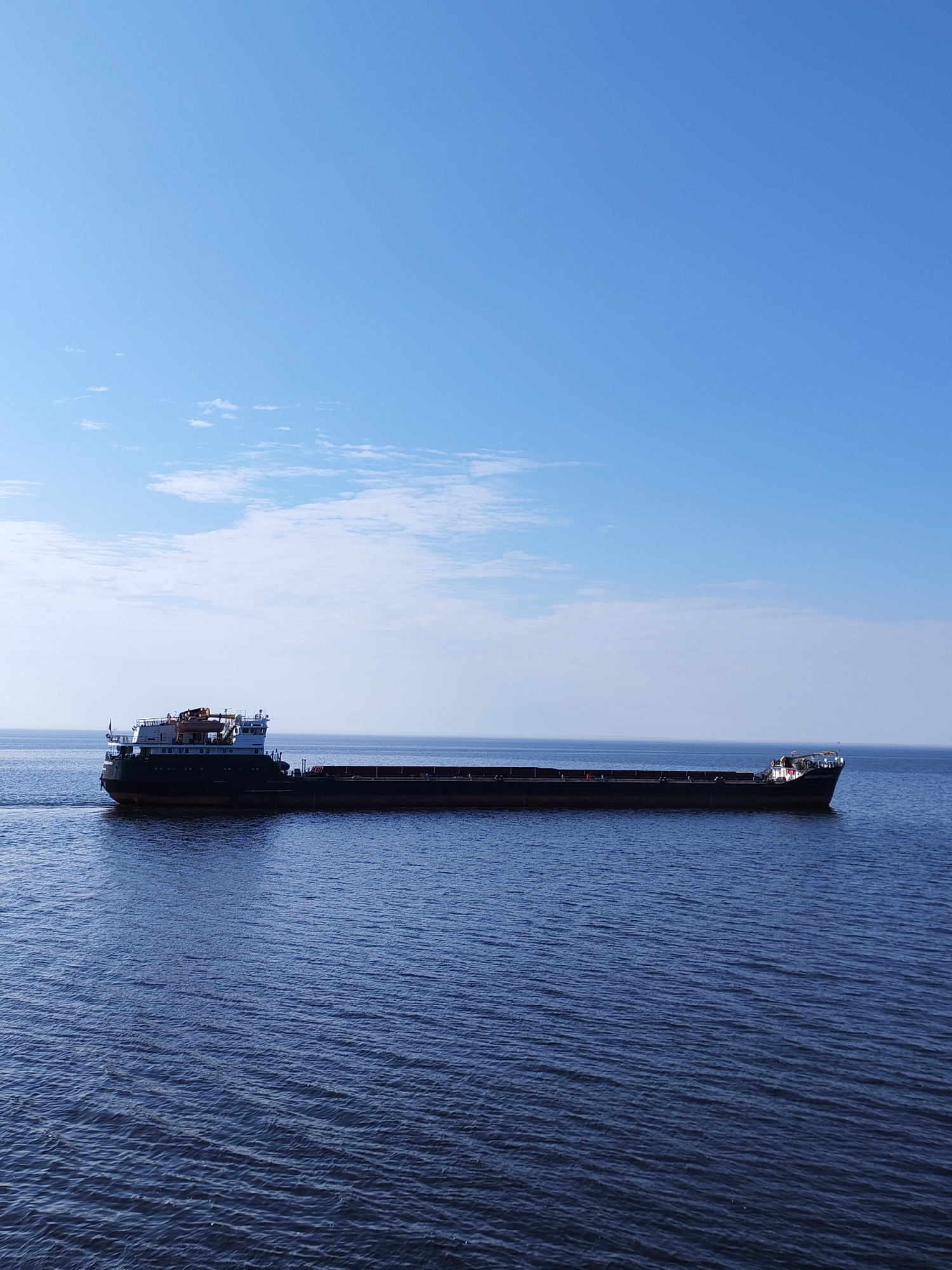 A long, empty barge traverses a body of water under a mostly clear, light blue sky with a few wispy clouds. The water is dark blue and slightly rippled. The barge is dark in color, with a raised section at the front of the vessel. The image does not contain any people. The location, Mologa, Russia, cannot be directly confirmed from visual elements in the image.