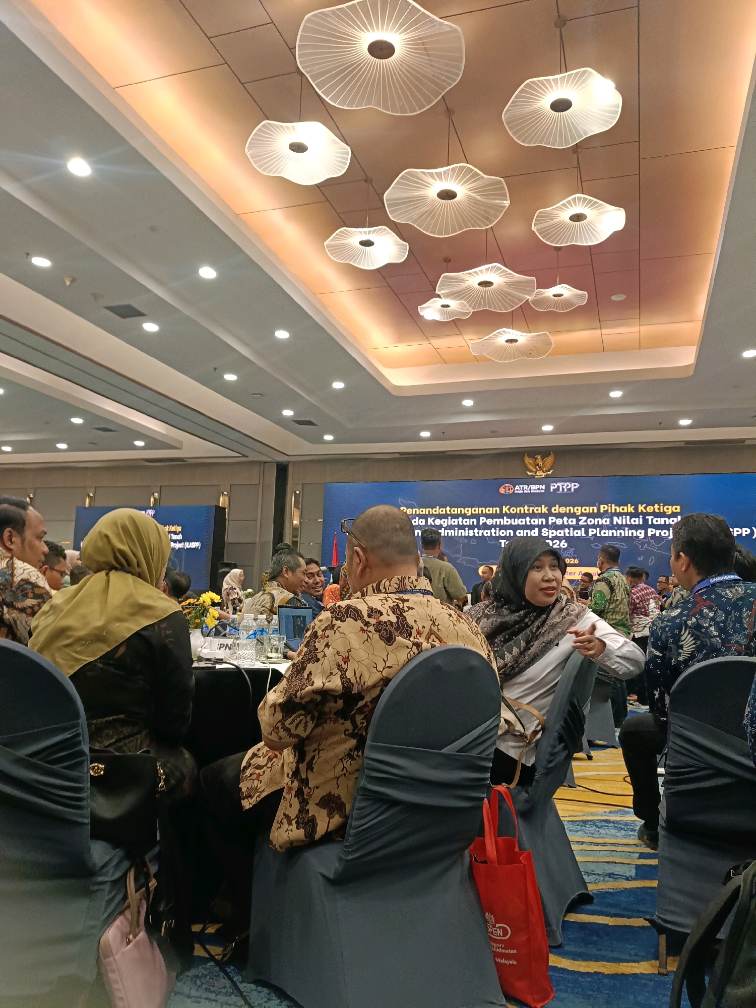 The image depicts an indoor event, likely a conference or seminar, with an audience seated at tables in a large room. The ceiling features decorative, cloud-shaped light fixtures and recessed lighting. A large banner at the front of the room displays text in Indonesian, including "Penandatanganan Kontrak dengan Pihak Ketiga" (Contract Signing with Third Parties) and "Kegiatan Pembuatan Peta Zona Nilai Tanah" (Land Value Zone Map Creation Activities), with the mention of "Administration and Spatial Planning Project (ILASPP)" and the year "2026". Several people are visible, many wearing batik clothing, indicating a formal or semi-formal gathering. Some individuals are actively engaged in conversation or listening, while others appear to be observing the proceedings. The tables are set with water bottles and papers. The presence of the Indonesian national emblem on the banner suggests an official government or organizational event. This event appears to be taking place in the city of North Jakarta, Indonesia, a common location for such conferences and exhibitions.