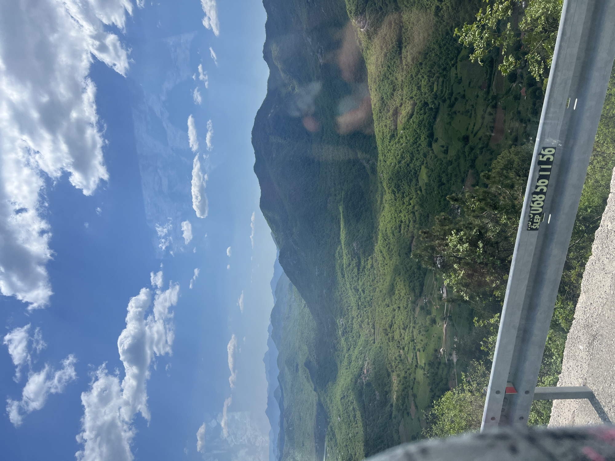 The image shows a daytime view of a mountainous landscape with a guardrail in the foreground. The sky is blue with scattered white clouds. The mountain is covered in lush green vegetation. The guardrail is gray and has a sticker with the phone number "SLEP U068 56 11 56" on it. The location is Dobrsko selo, Montenegro.