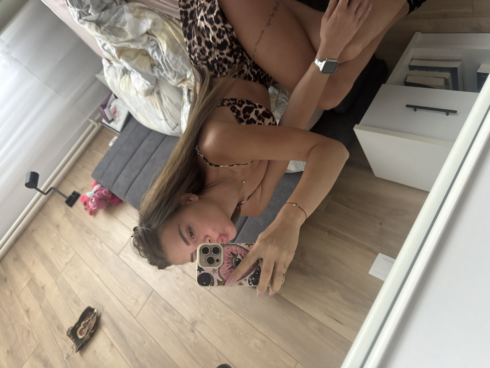 A woman is lying on a bed, taking a selfie with her phone. She is wearing a leopard-print top and matching bikini bottoms. The bed has a white comforter and gray headboard. A white nightstand with an open drawer and books is on the right side of the bed. The floor is wood. A black desk lamp and pink stuffed animal are near the floor on the left. A shoe or slipper is also on the floor. A white-framed mirror is on the right side of the image, reflecting the scene. The location appears to be a bedroom.